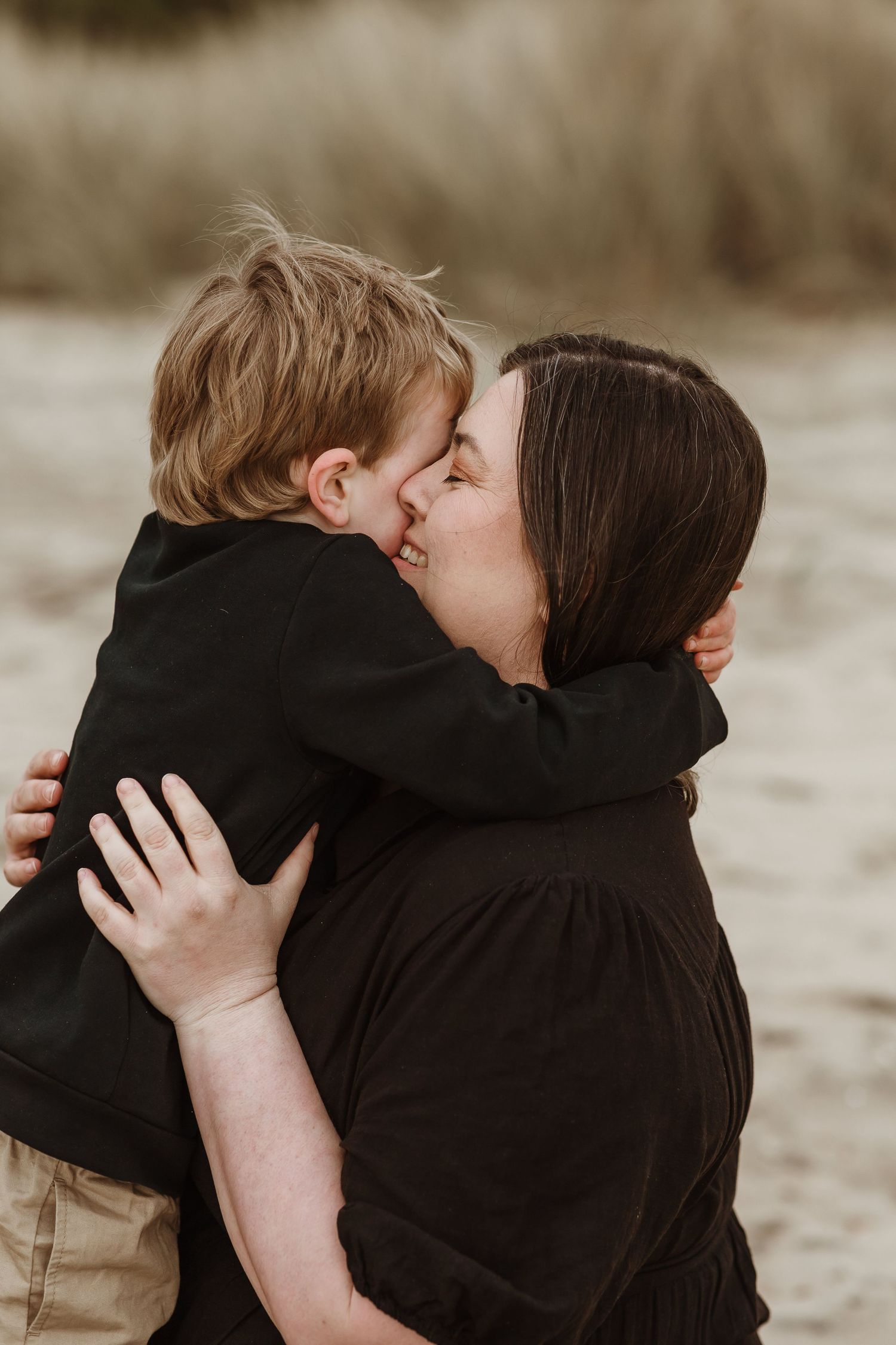 A mother tenderly kisses her young son on a sandy beach, both dressed in black, sharing a loving embrace.