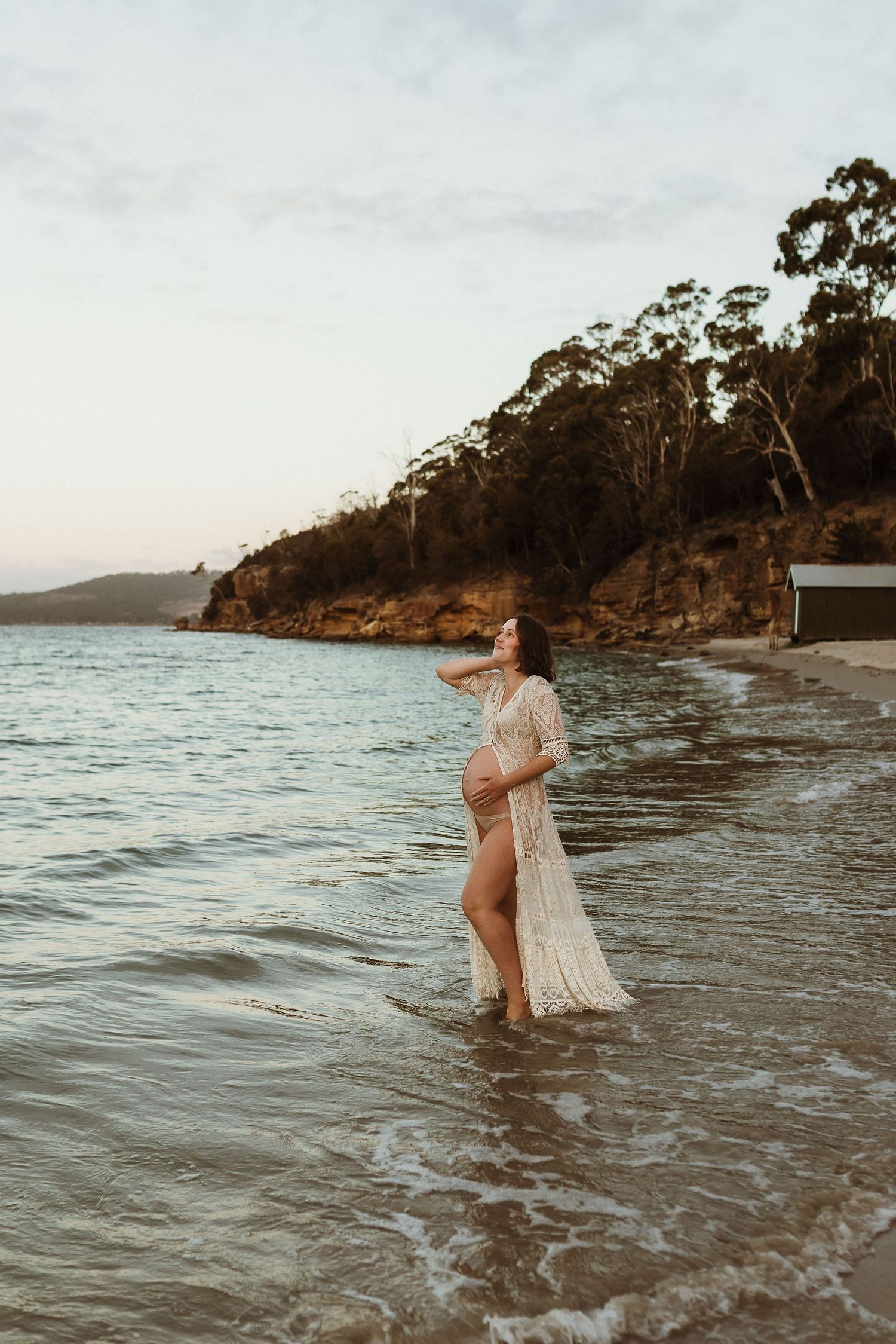 Pregnant woman in lace dress standing in ocean waves on a serene beach at sunset.