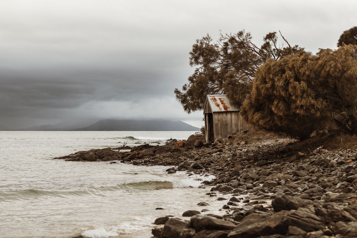 Moody coastal landscape with small wooden structure perched on rocky shore at sunset.