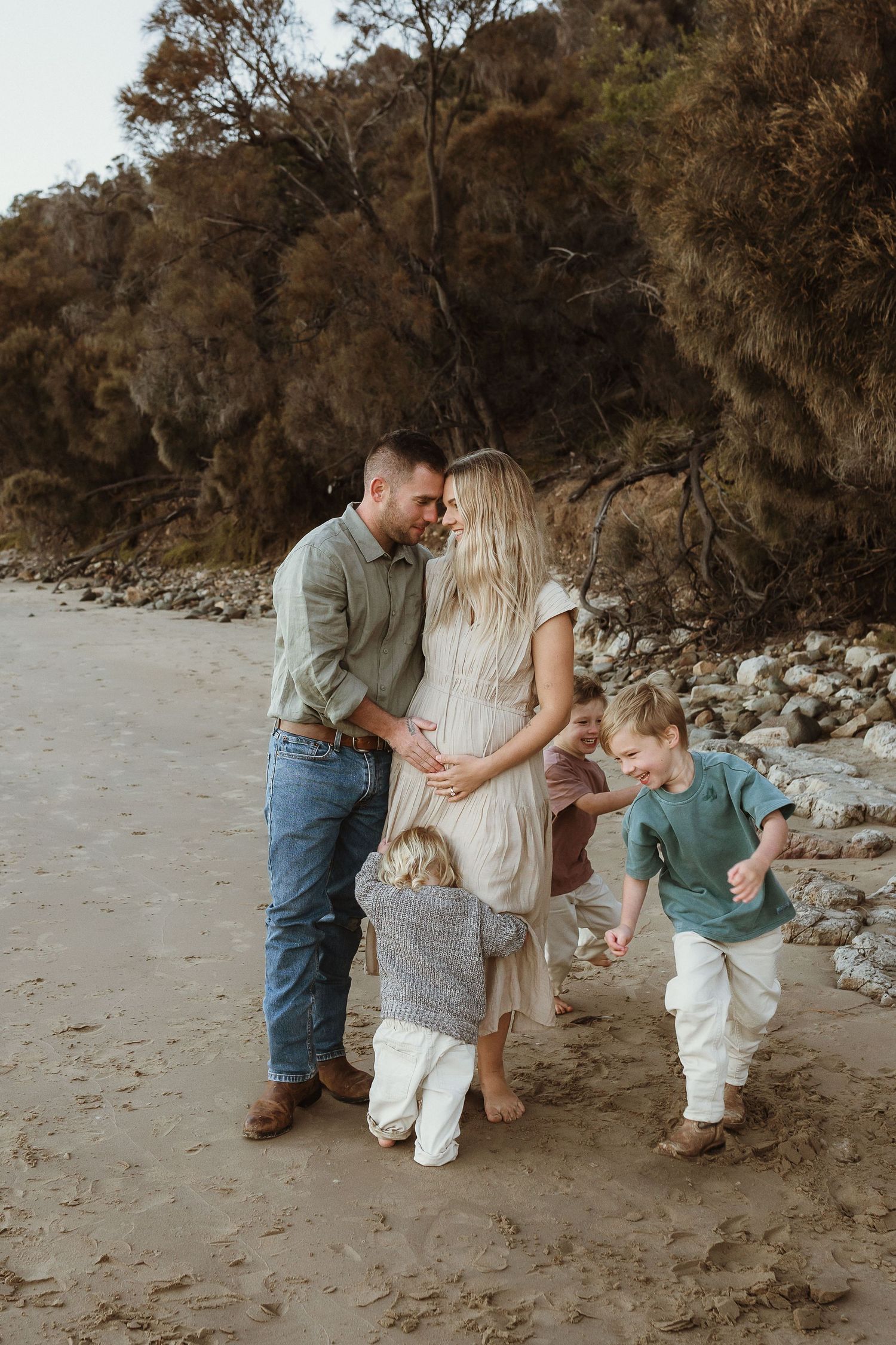Pregnant woman and man share a tender moment on a sandy beach while three young children play around them.