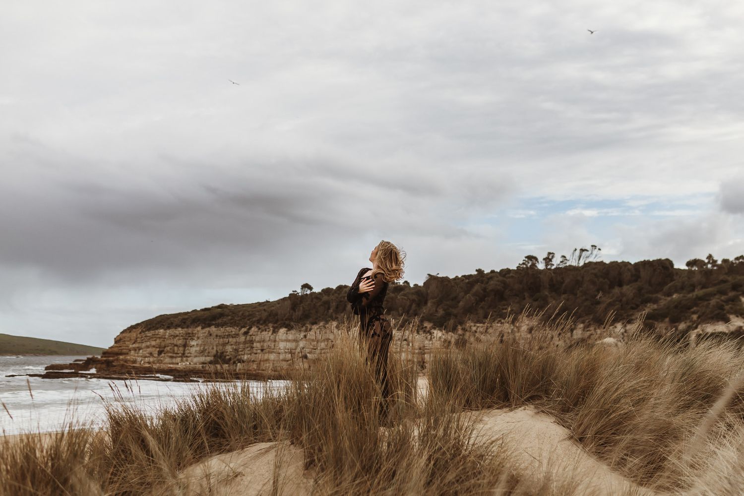 Figure stands among coastal grass dunes overlooking dramatic ocean cliffs.
