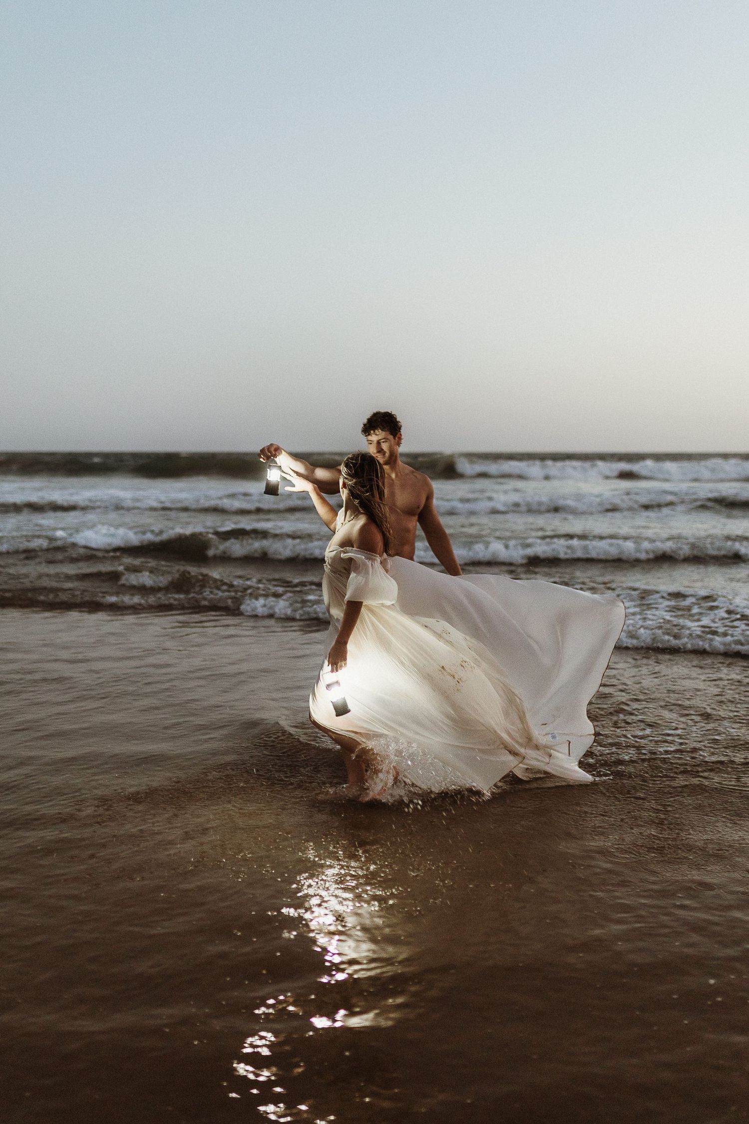 Bride and groom dancing in ocean waves at sunset, white wedding dress flowing in the breeze on a sandy beach.