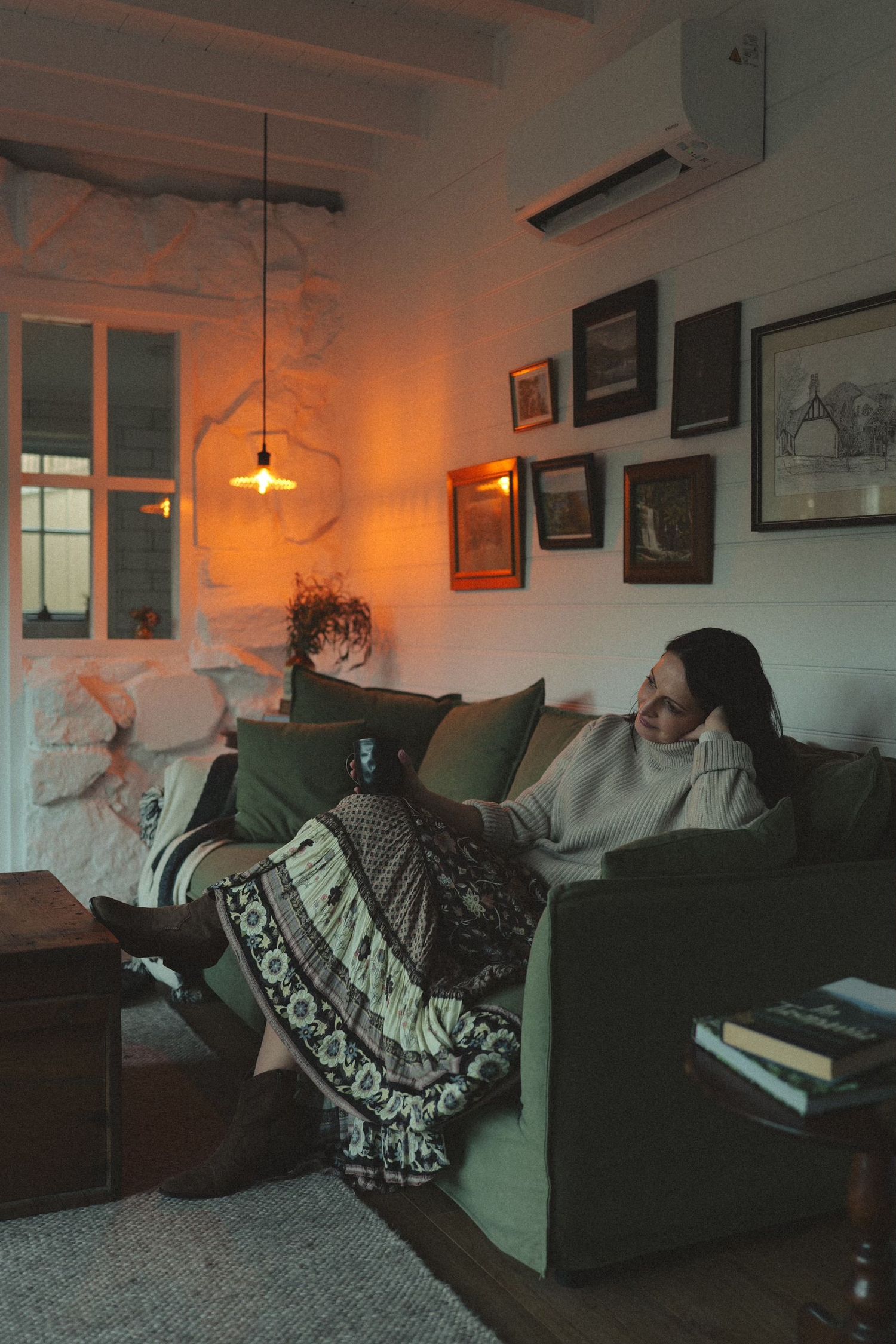 Woman relaxing on green sofa in cozy warmly lit living room with gallery wall and bohemian decor.