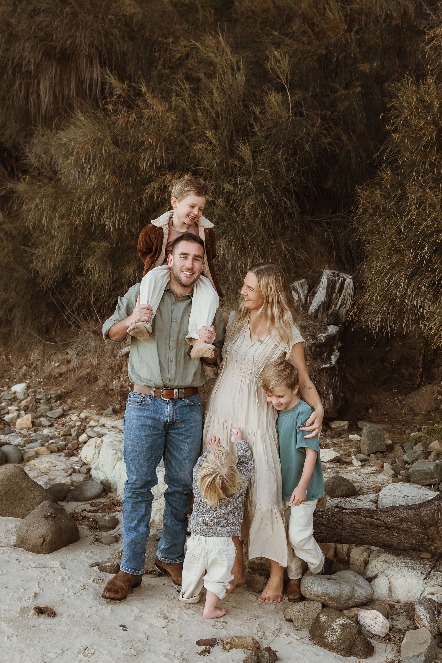 Happy family of five posing outdoors on rocky beach with lush greenery backdrop during casual photo session.