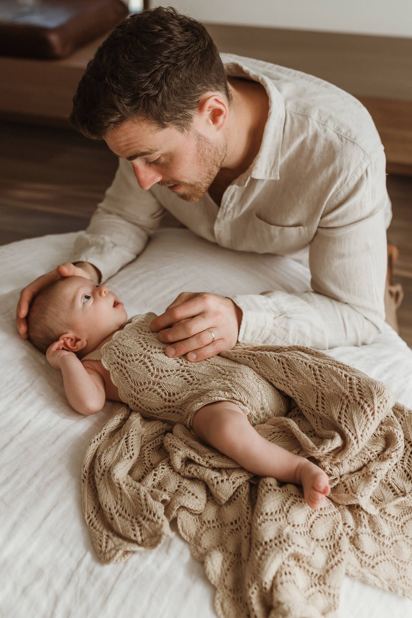 A tender moment between parent and baby lying on white bedding wearing a knit dress.