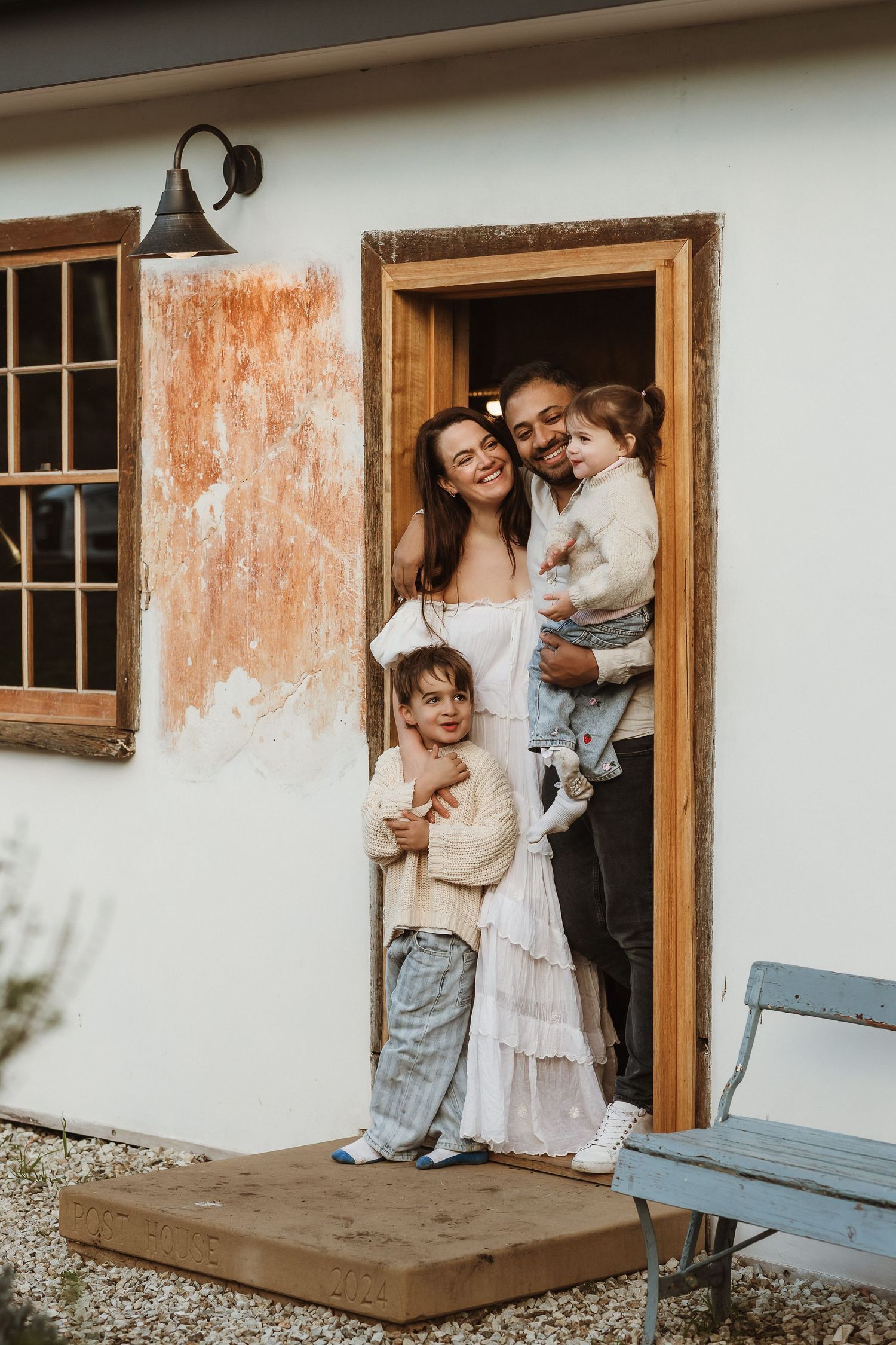 A casual couple embraces playfully against a rustic white wall with peeling paint and wooden window frames.