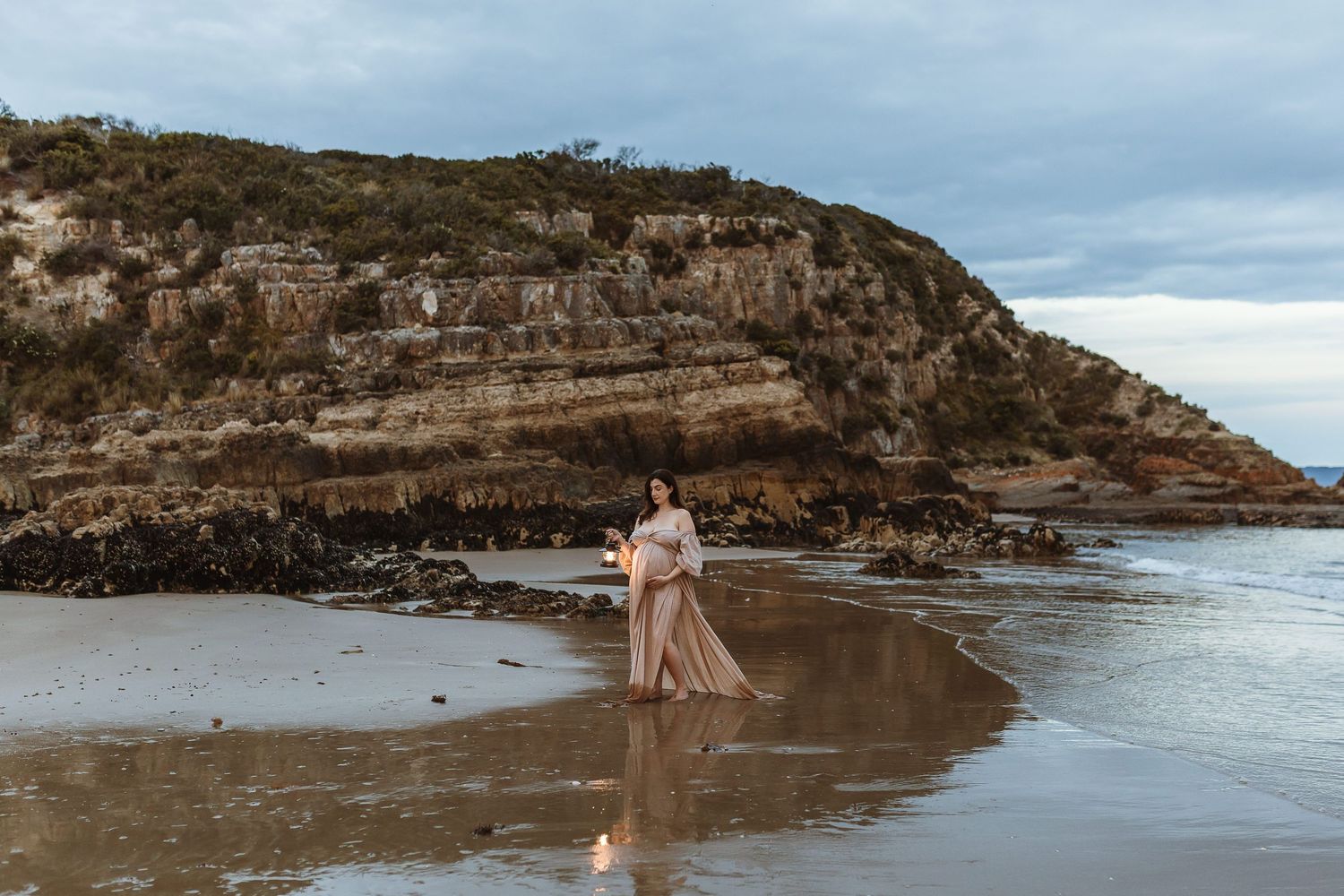 Pregnant woman in flowing gown stands on sandy beach with rocky cliffs backdrop at dusk.