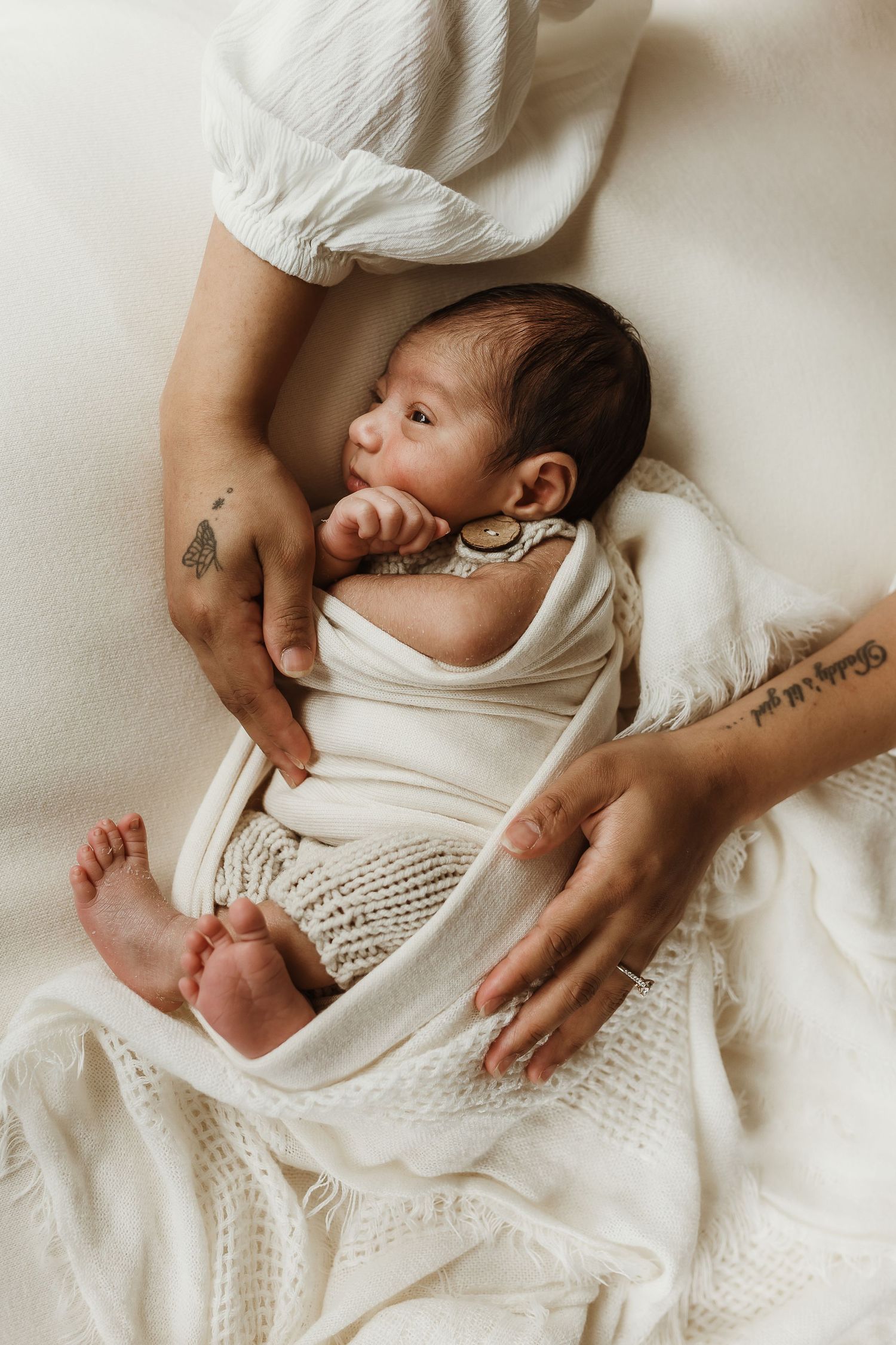 Newborn baby swaddled in white blanket cradled by tattooed mother's hands on soft white bedding.