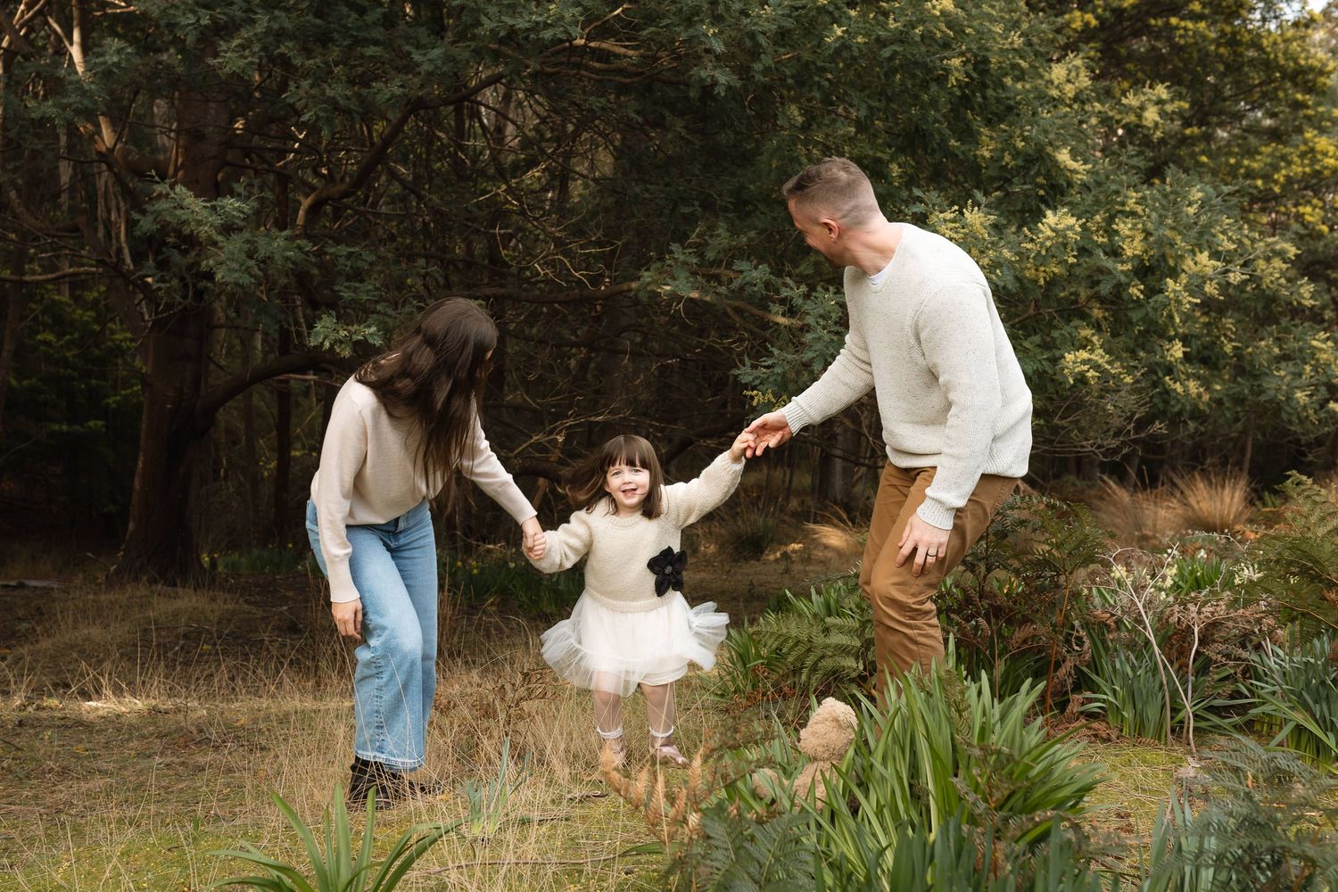 A family of three holds hands in a lush garden, parents swinging their young daughter in a white dress.