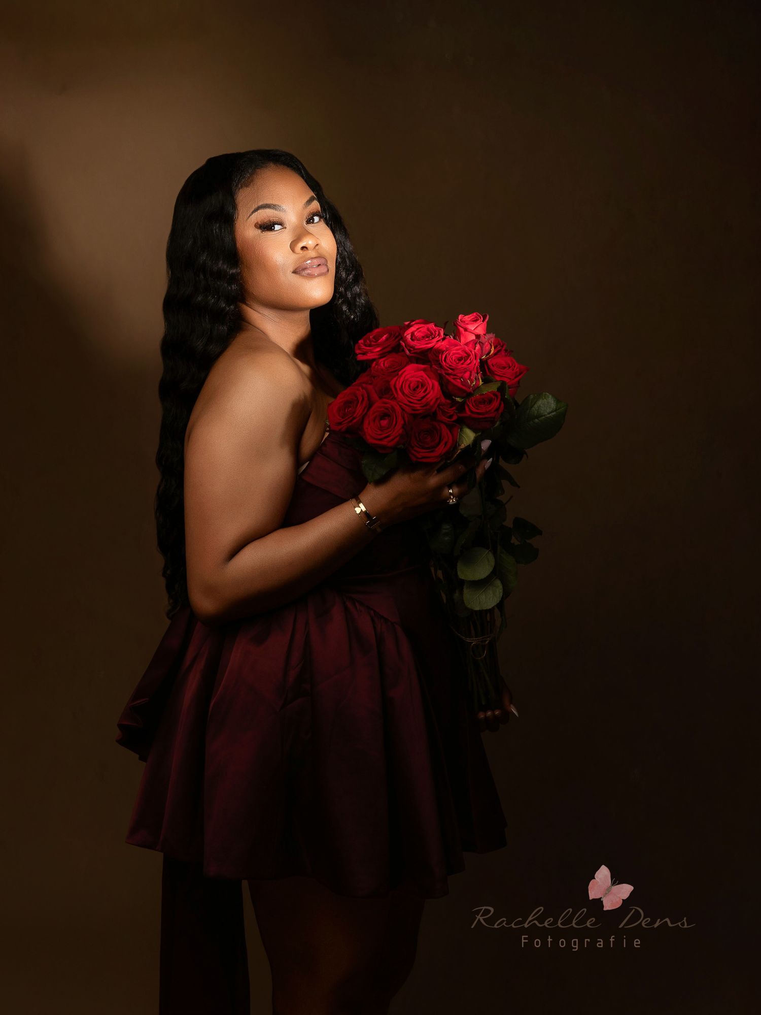 A woman in a burgundy dress poses elegantly with red roses against a dark studio backdrop in a glamour photoshoot.