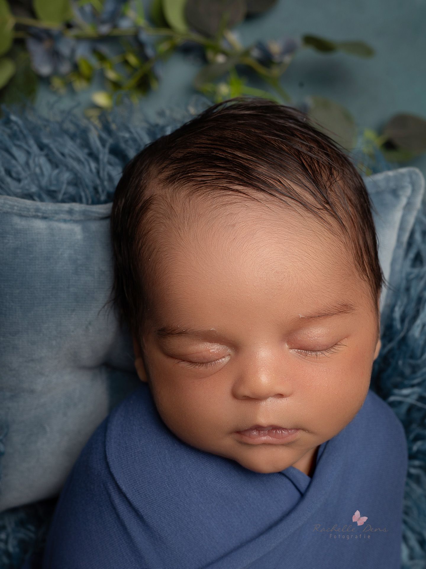 A peaceful sleeping newborn wrapped in soft blue fabric rests on light blue bedding with green foliage in the background.