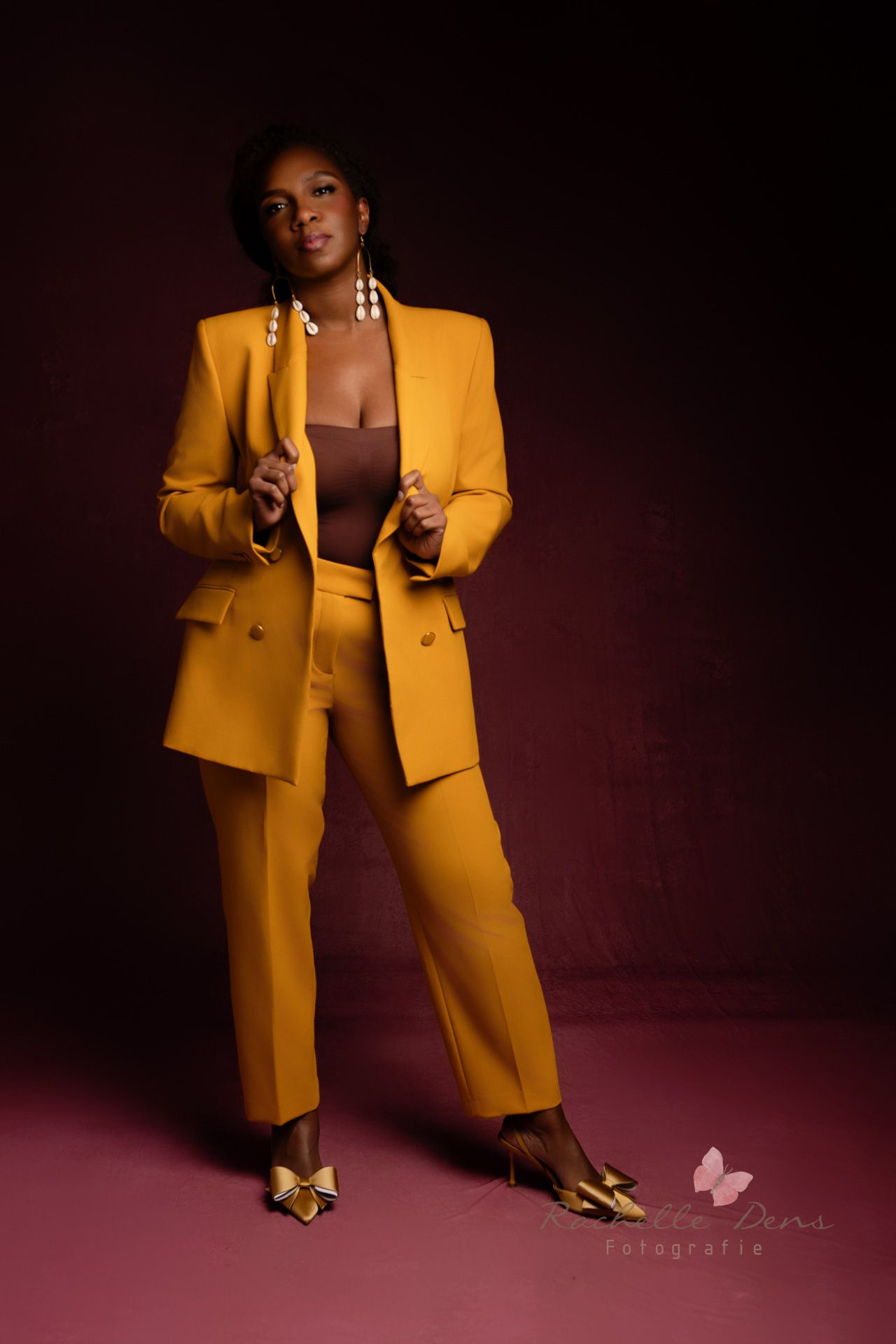 A stylish yellow power suit ensemble with gold heels photographed against a dark burgundy background in a studio setting.