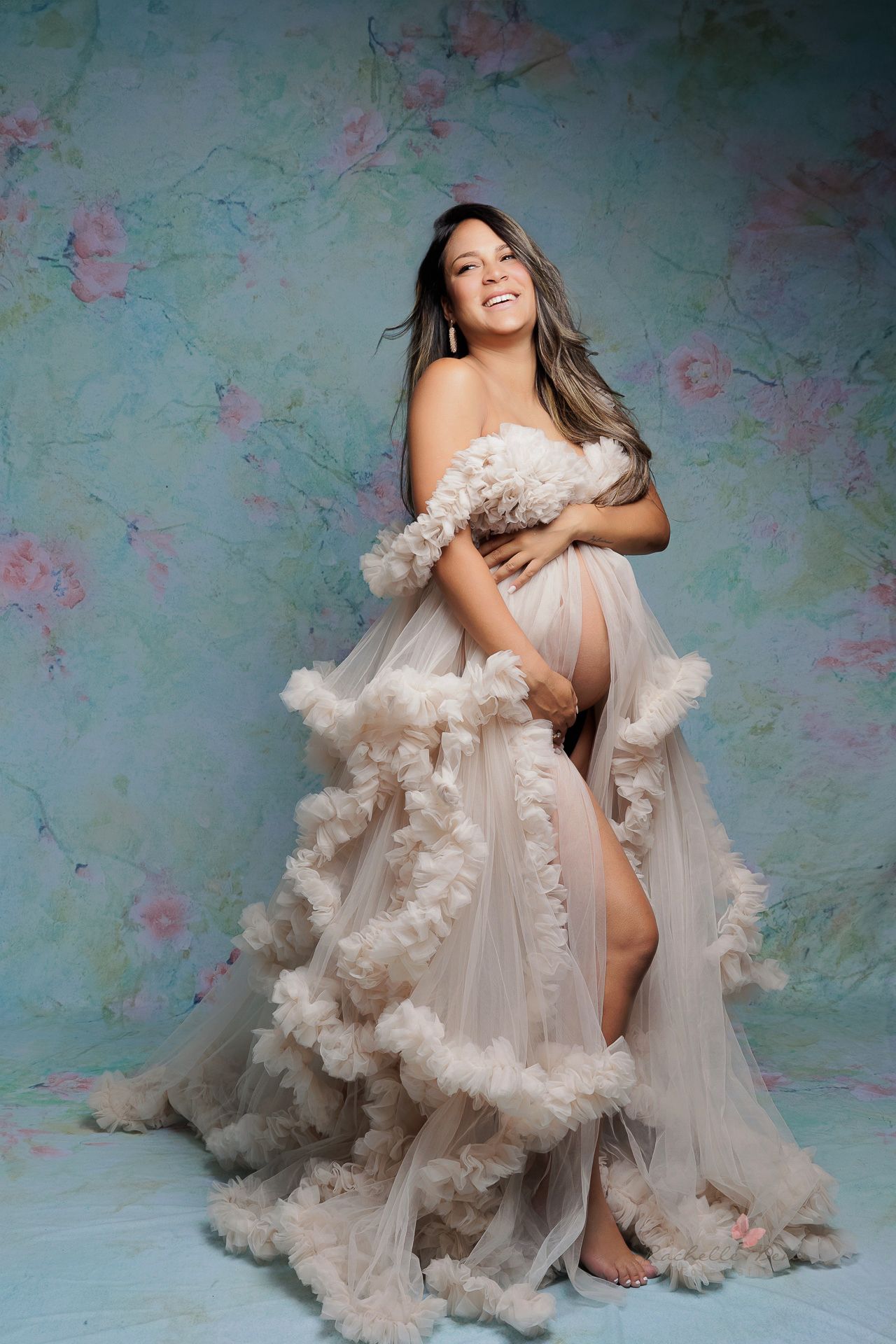A dreamy maternity photoshoot featuring an ethereal white tulle gown with floral appliques against a blue floral backdrop.