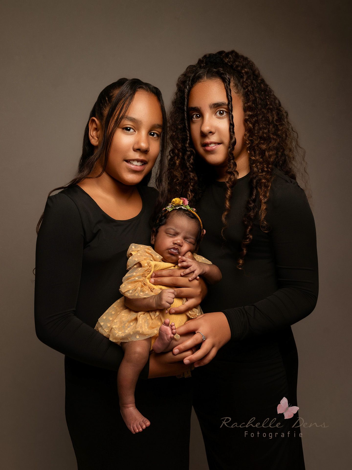 Studio portrait of two people in black tops holding a baby wearing a gold dress against a dark background.
