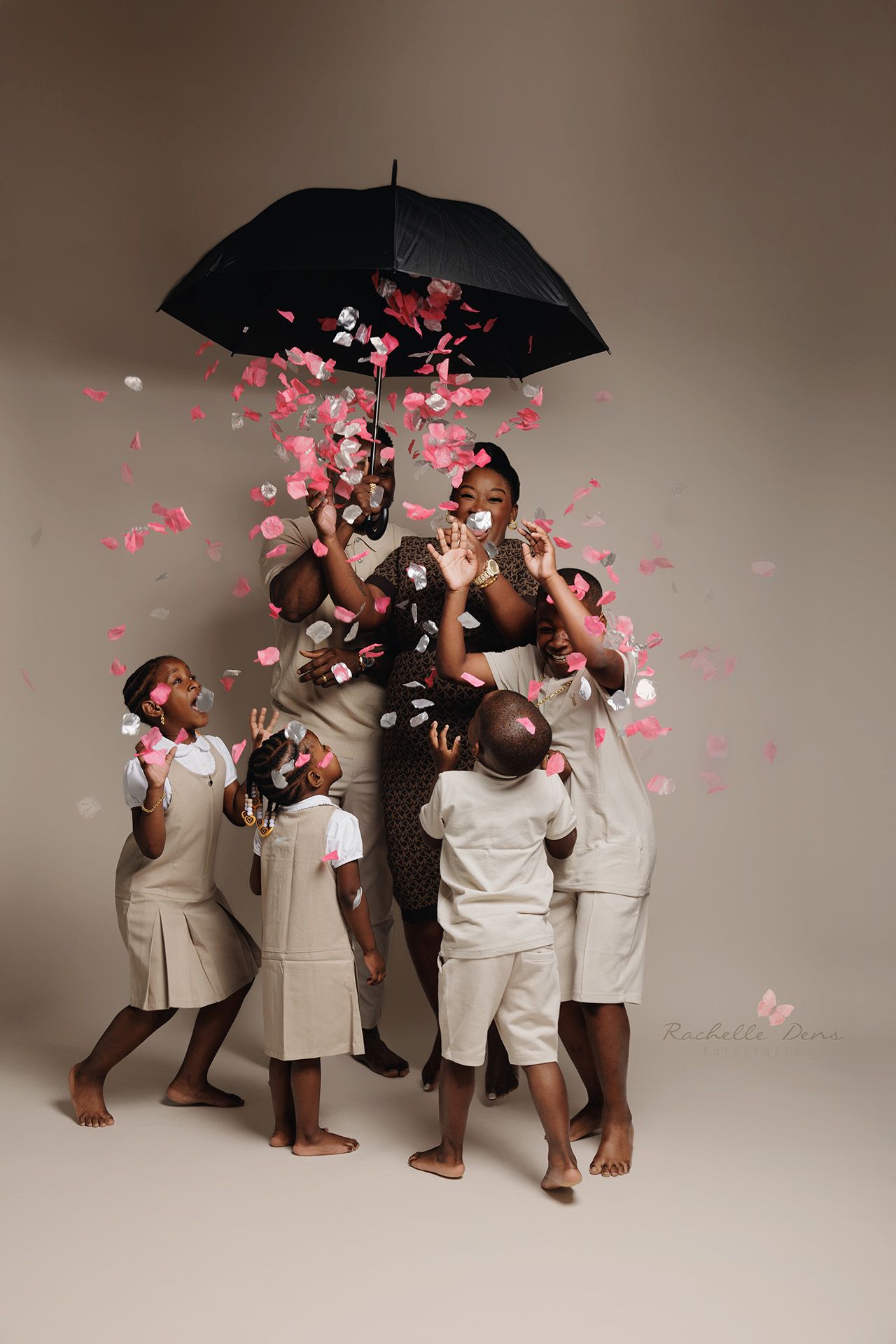 A group in white outfits playfully gathers under a black umbrella while pink rose petals fall around them in a studio setting.