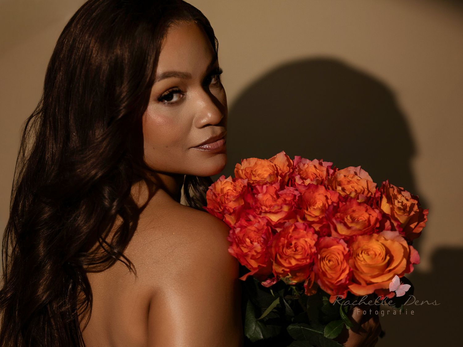 A woman in dark hair poses with vibrant orange roses against a neutral background.