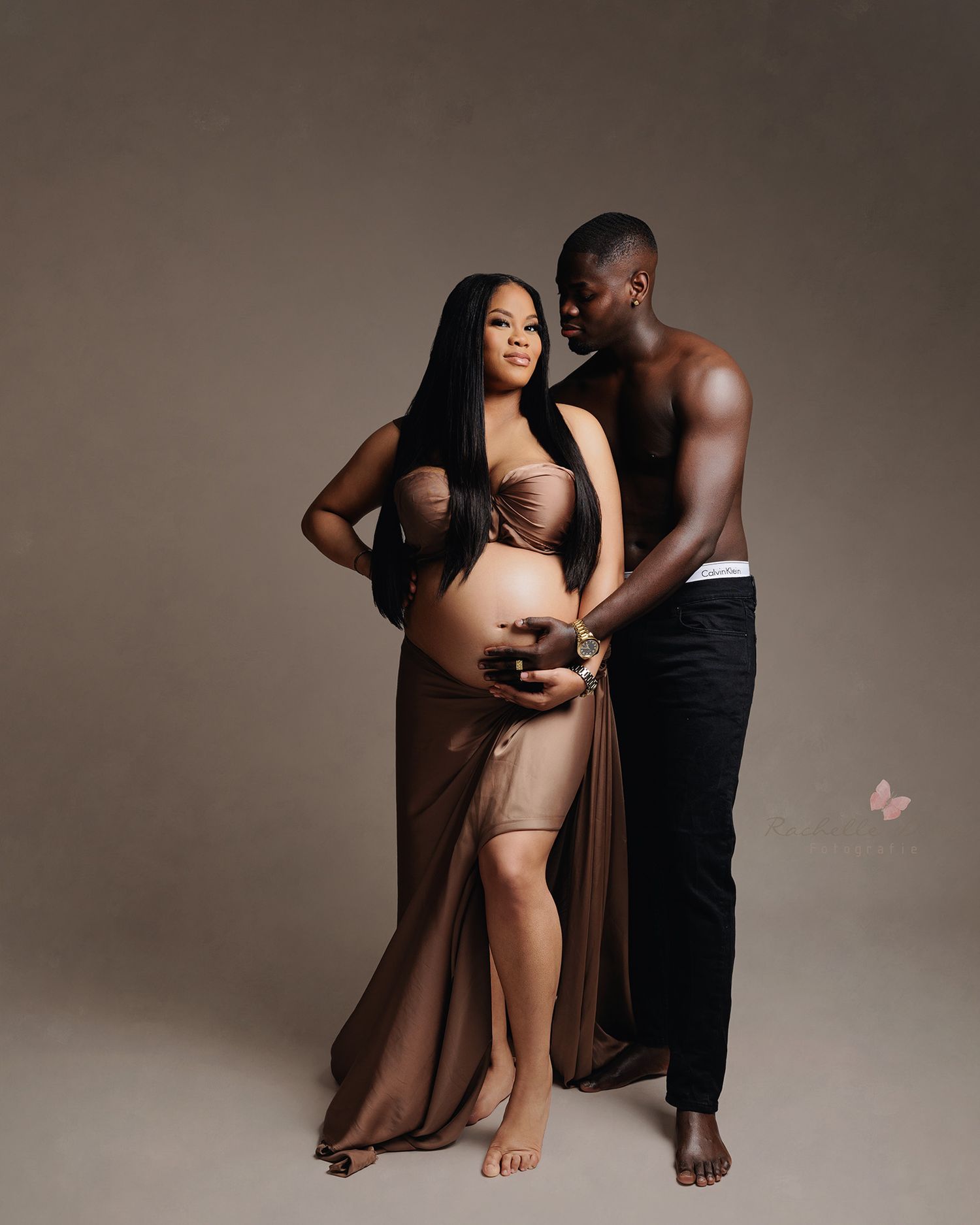 A romantic maternity photo of an expecting couple embracing in brown tones against a neutral studio backdrop.