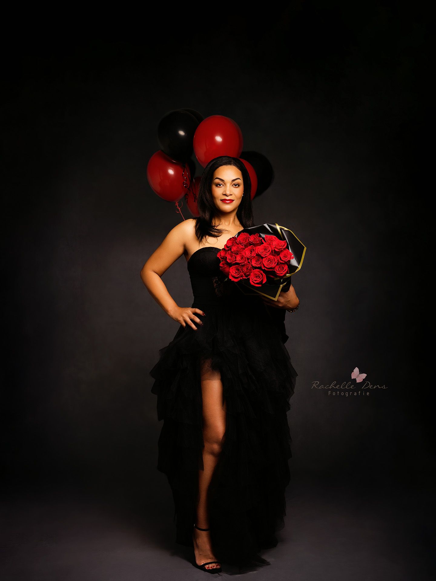Dramatic portrait with red balloons and roses against dark background featuring elegant black ruffled gown and glamorous styling.