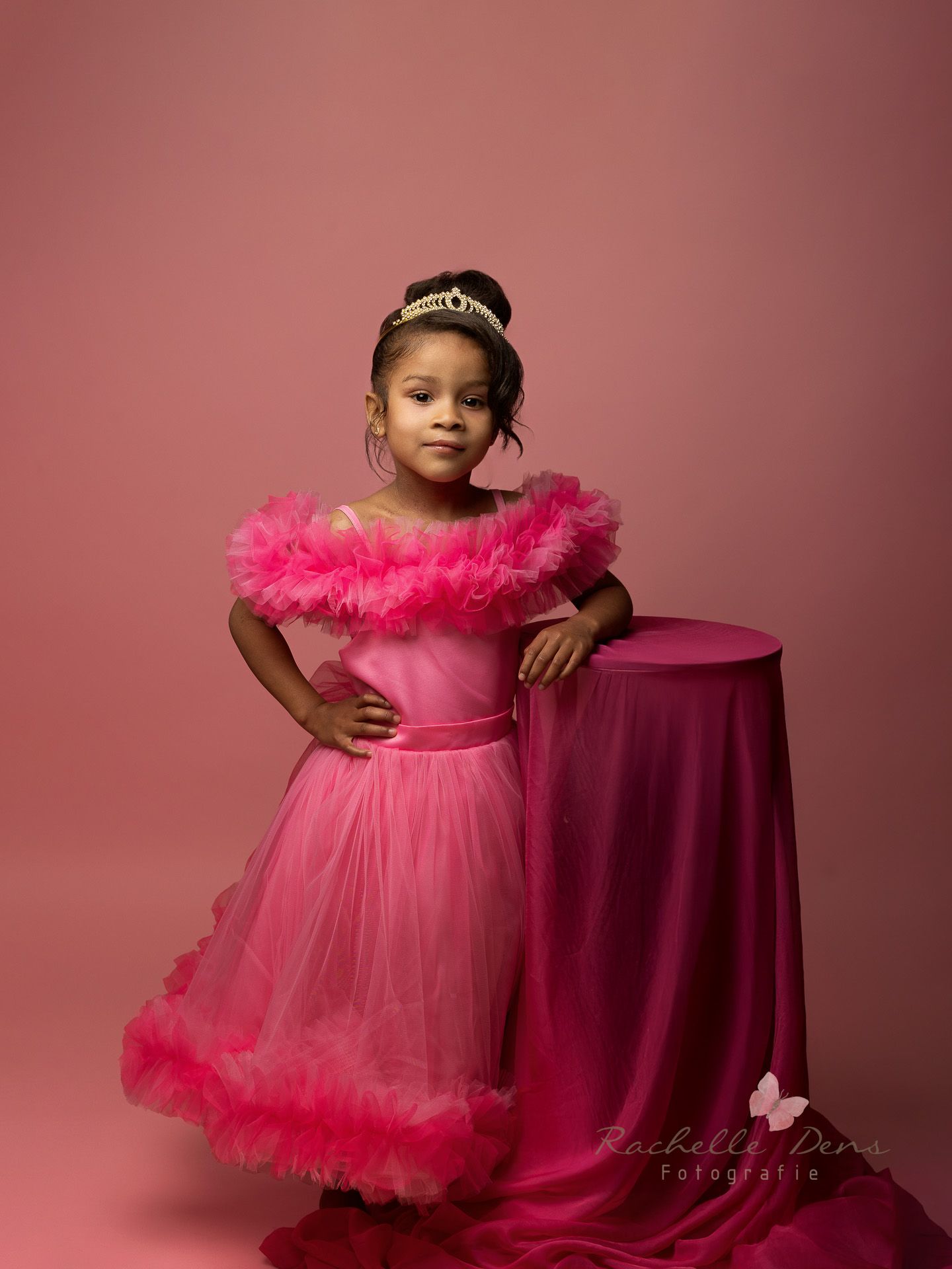 A child wearing a pink tulle dress with ruffled neckline poses next to a burgundy table against a rose-colored backdrop.