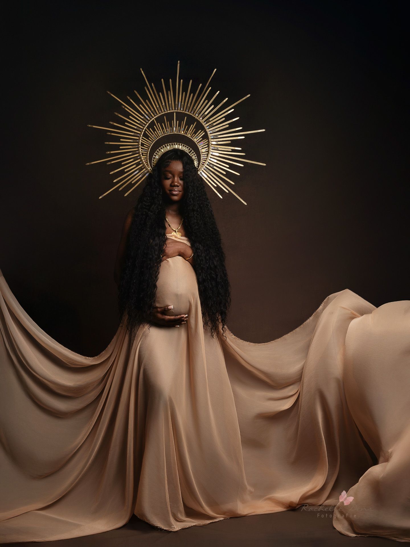 Artistic maternity portrait with flowing beige fabric and golden sunburst crown against dark background.