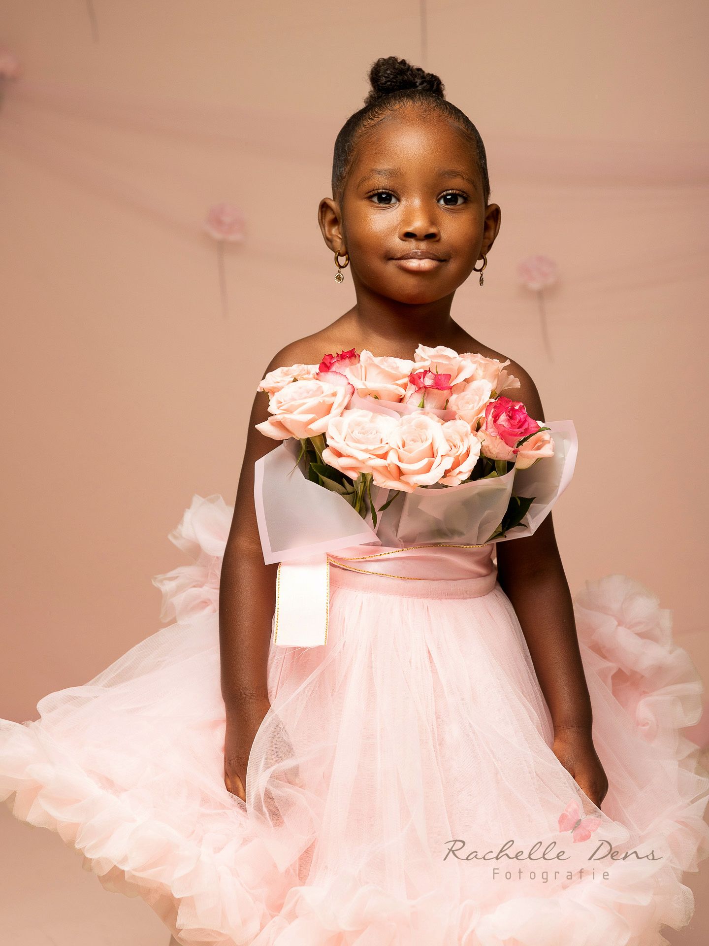 A child in a light pink tulle dress with ruffled neckline poses gracefully against a soft pink backdrop.