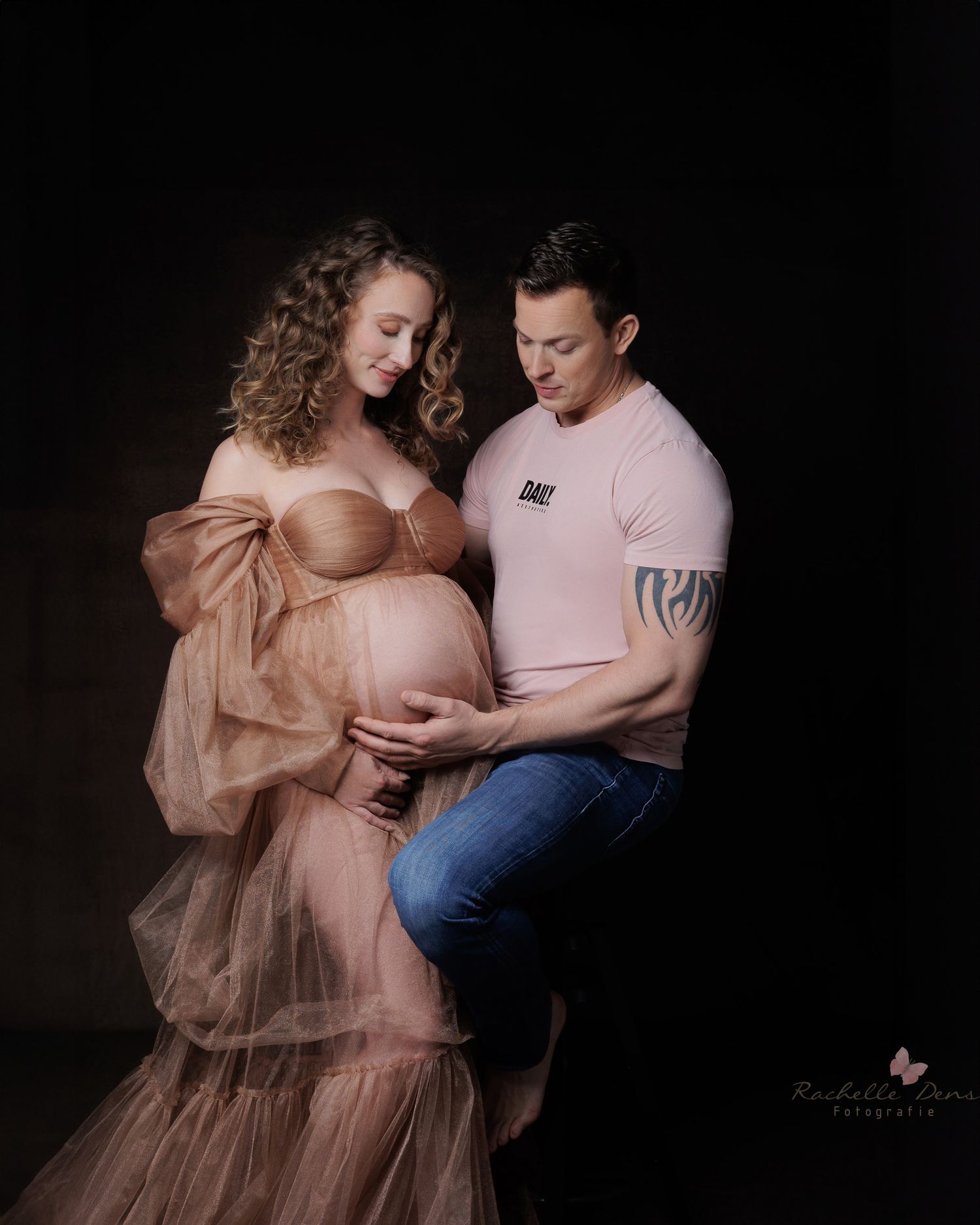 Romantic maternity photo in pink featuring a flowing tulle dress against dark background with couple embracing.