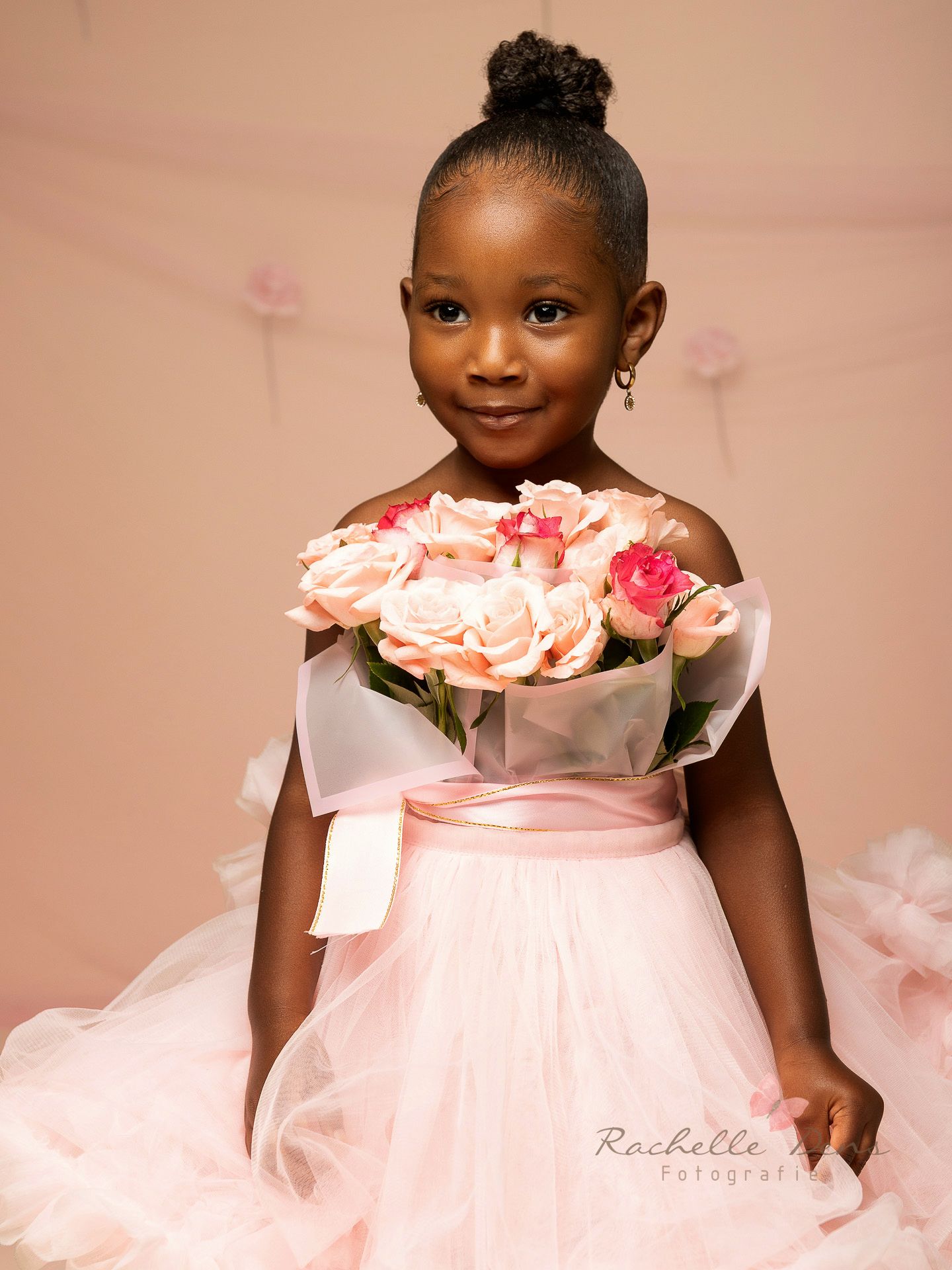 Portrait of a child in a pink tulle dress holding a bouquet of pink roses against a blush-colored backdrop.