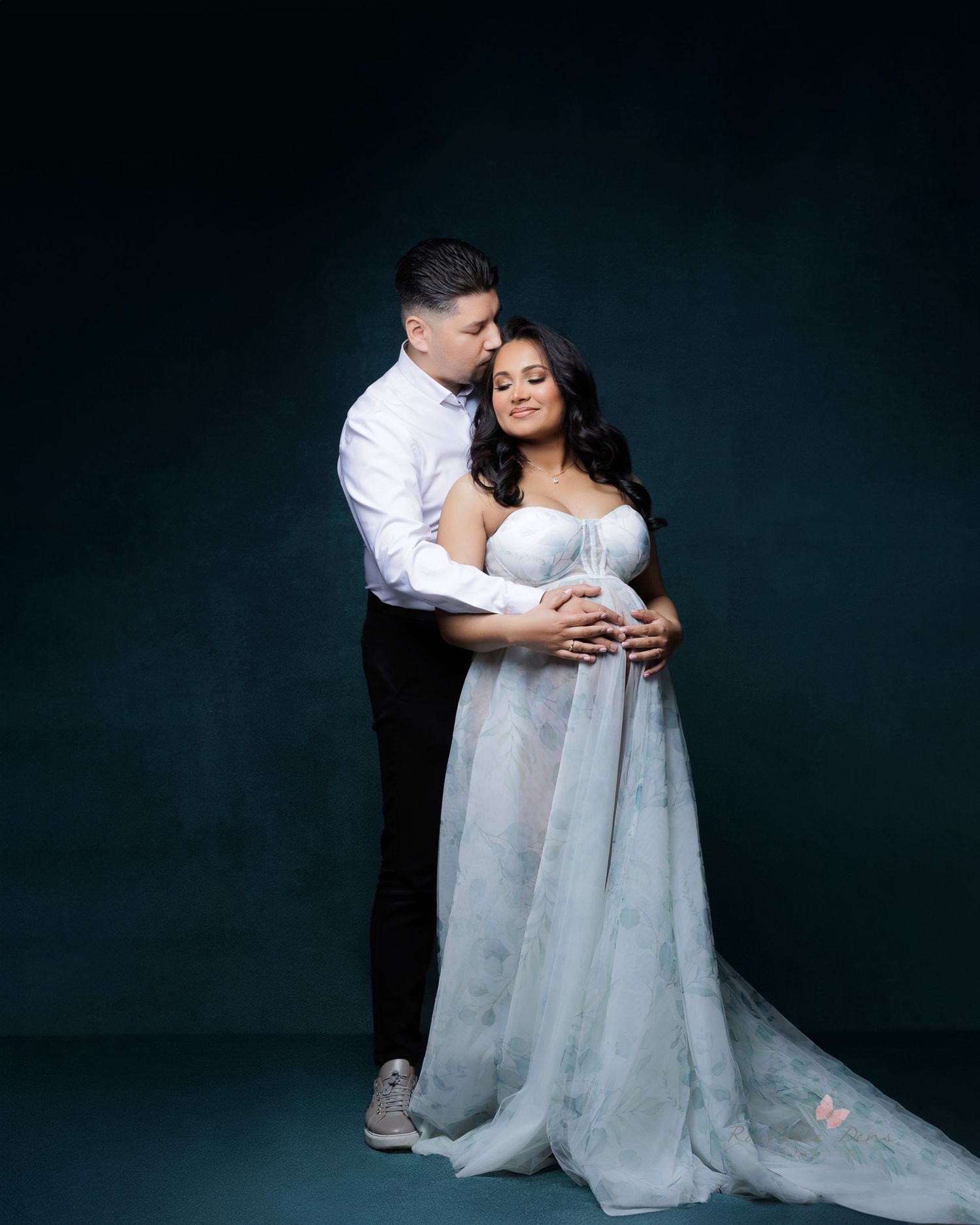A couple in white attire share an intimate moment during a romantic photo session against a dark backdrop.