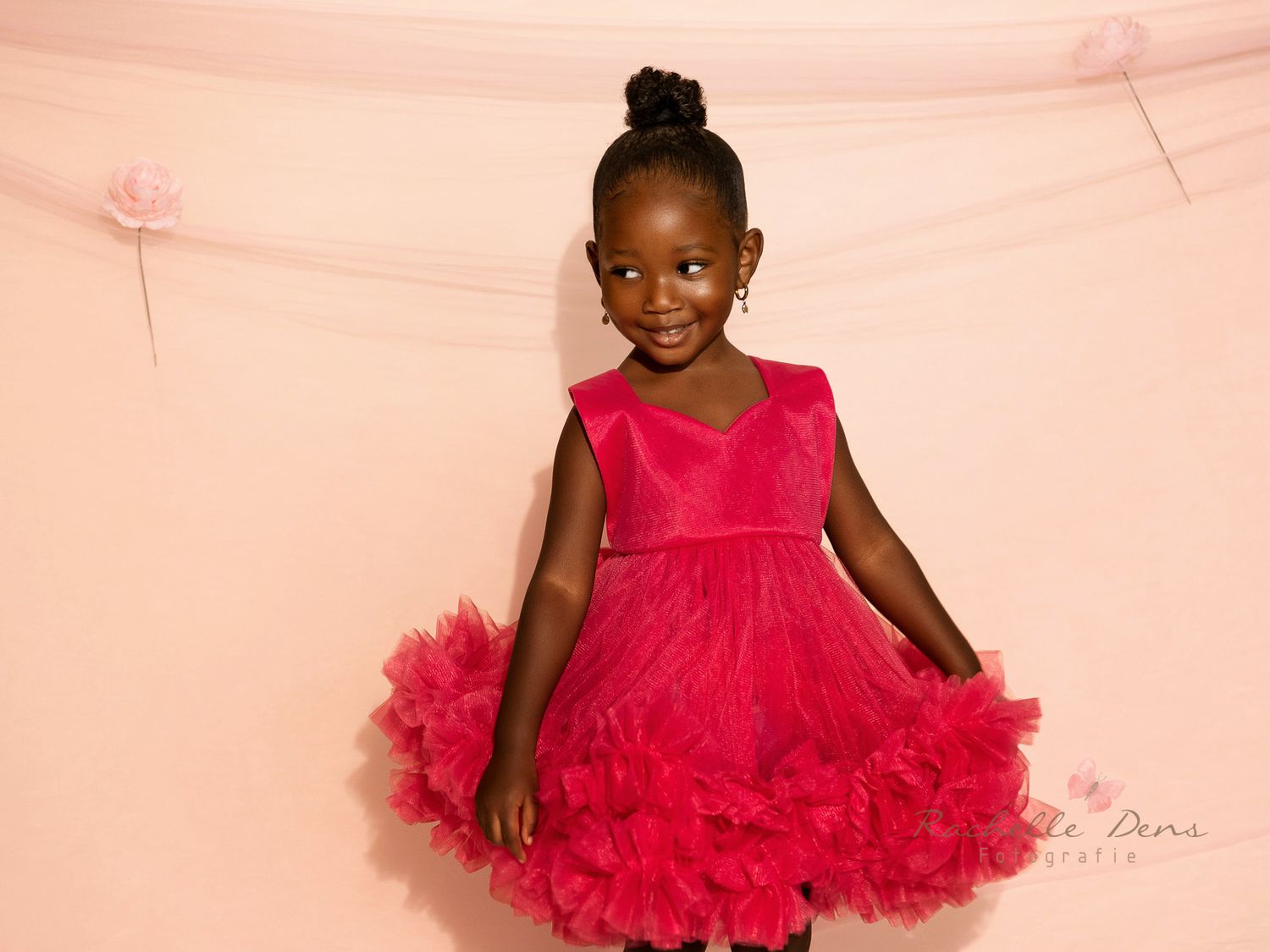 A young child twirls in a vibrant red tulle dress with ruffles against a soft pink background.
