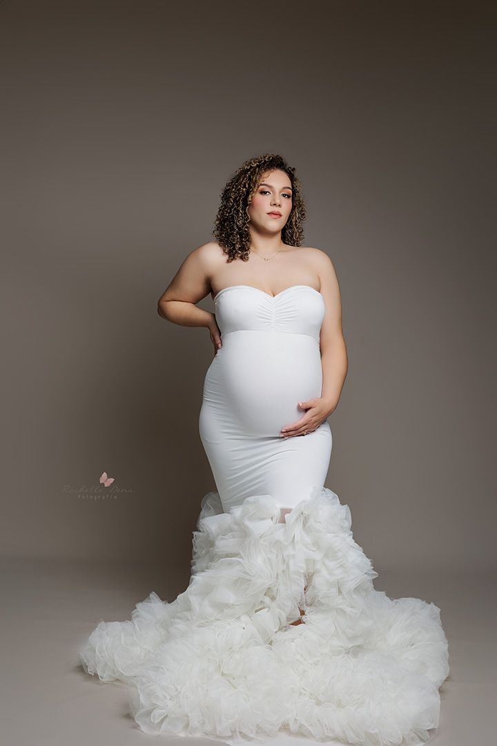 Expectant mother poses in elegant white mermaid-style wedding gown with dramatic ruffled train against neutral backdrop.