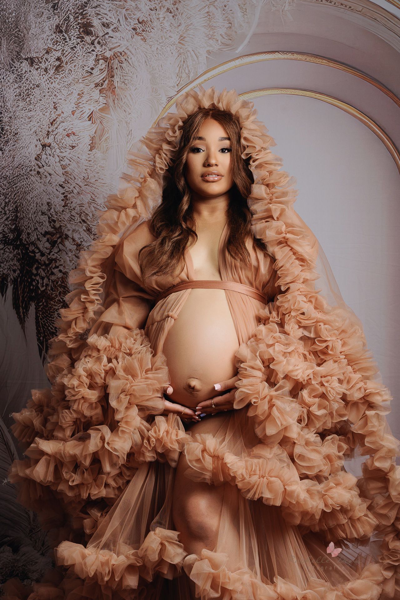 Ethereal maternity portrait featuring a flowing peach-colored ruffled tulle gown against an arched textured backdrop.