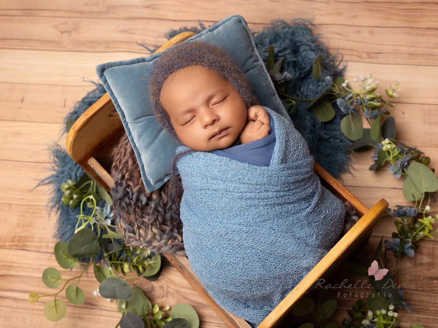 Sleeping newborn wrapped in blue swaddle blanket lying on wooden floor with greenery and floral decorations.