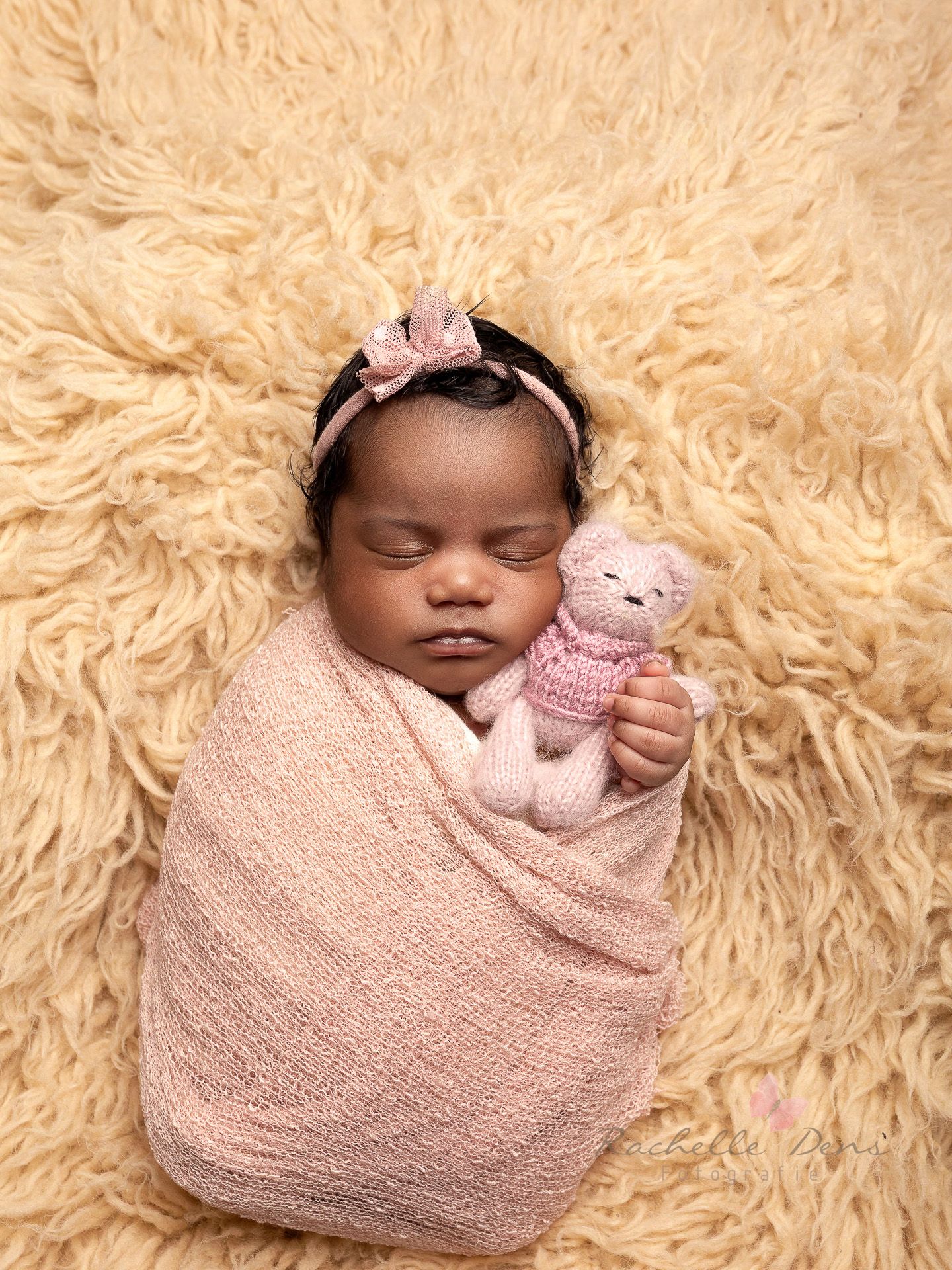 Sleeping baby wrapped in pink swaddle with matching headband and teddy bear on fluffy cream-colored fur background.
