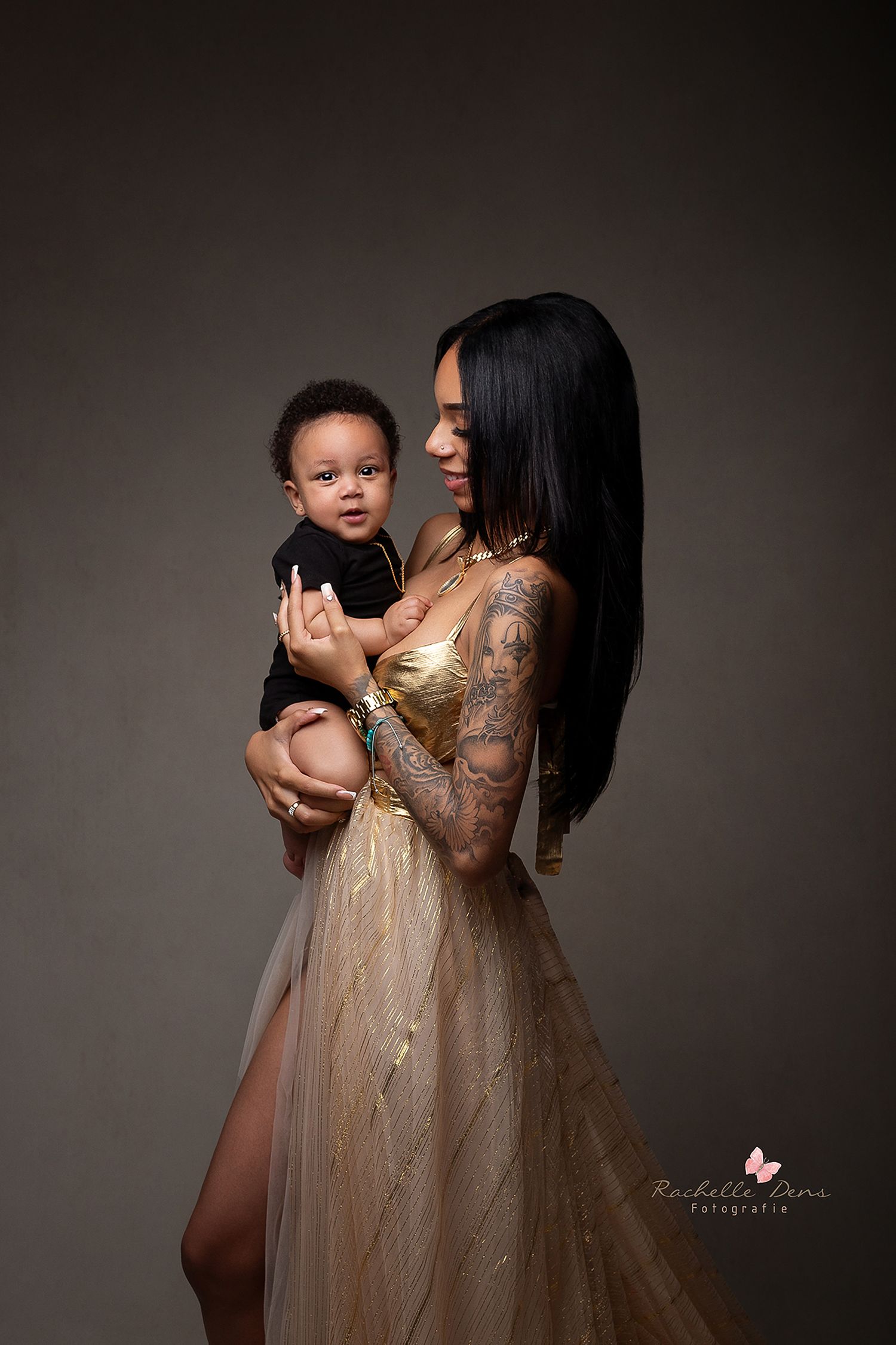 An intimate studio portrait showing parent and child in elegant gold dress against dark background.