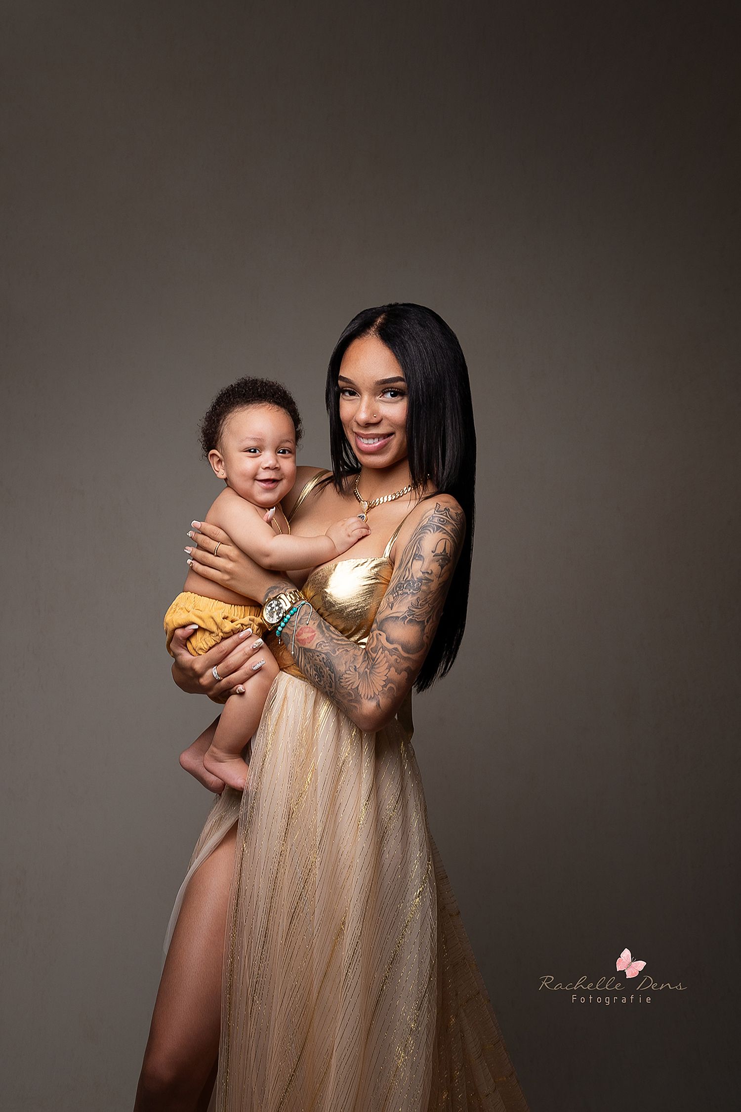 Elegant studio portrait of parent and toddler in gold flowing dress with tattoo sleeve against grey background.