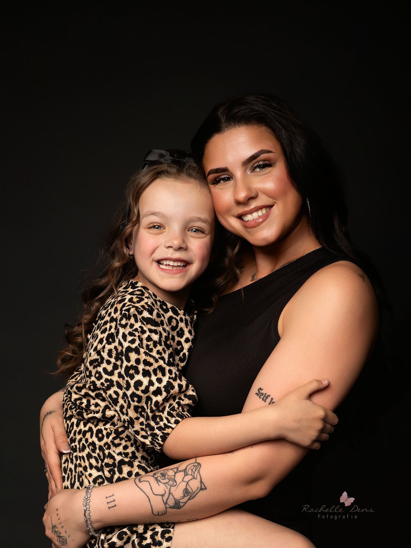 Portrait of two people in black and leopard print against dark background, sharing warm smiles and an embrace.