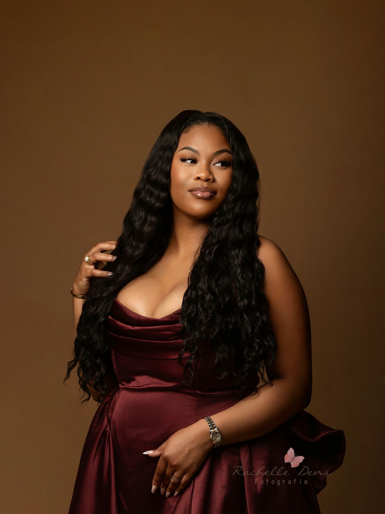 A portrait of someone wearing a burgundy satin gown against a brown backdrop with long flowing dark hair.