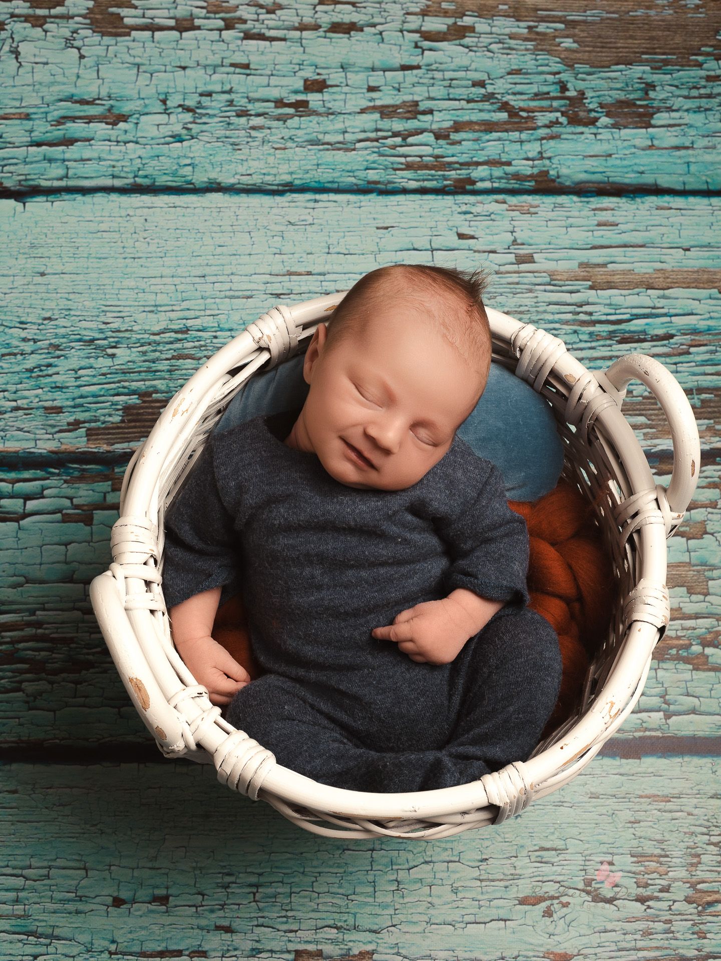 Baby sleeping peacefully in a vintage white wicker basket against distressed turquoise wooden backdrop.