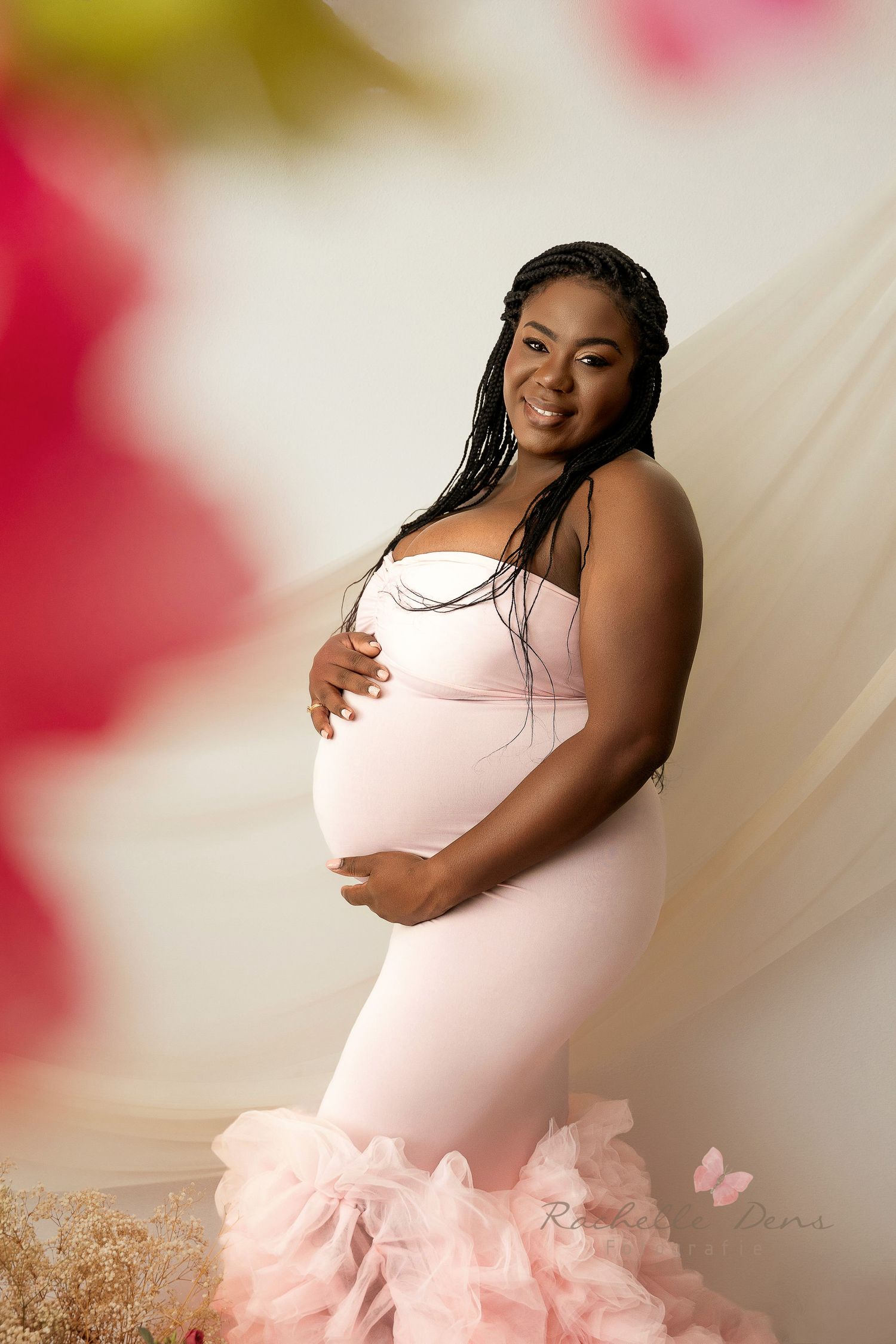 A pregnant woman poses in a blush pink mermaid-style gown with ruffled tulle details against a soft dreamy background.