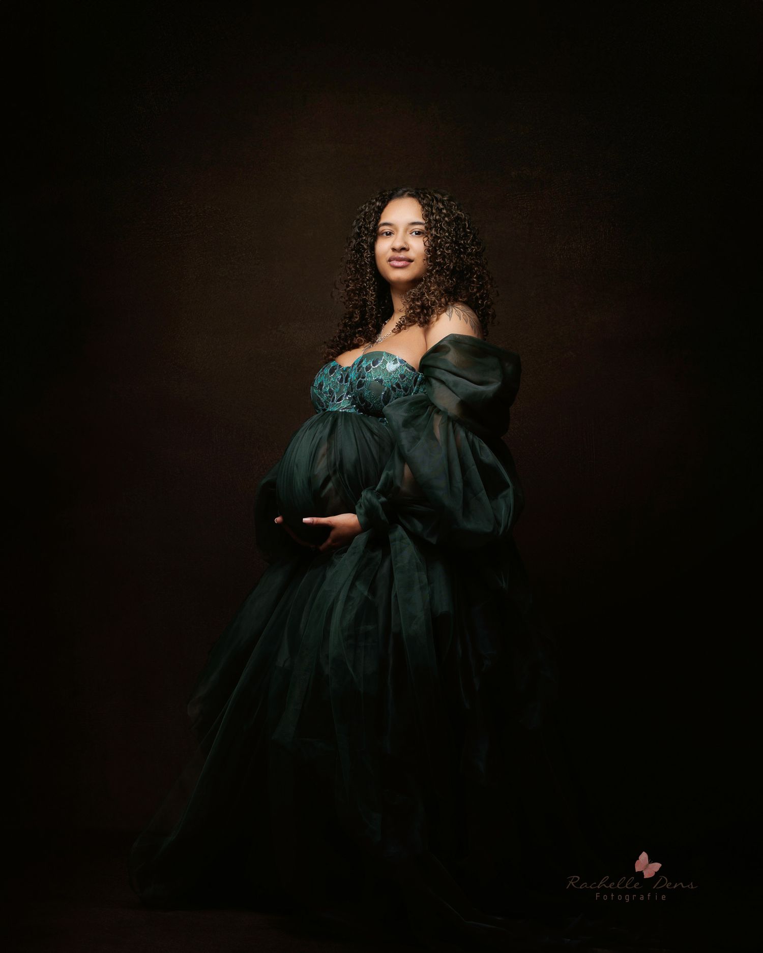 A maternity portrait in a dark emerald green Renaissance-style gown against a moody brown background.