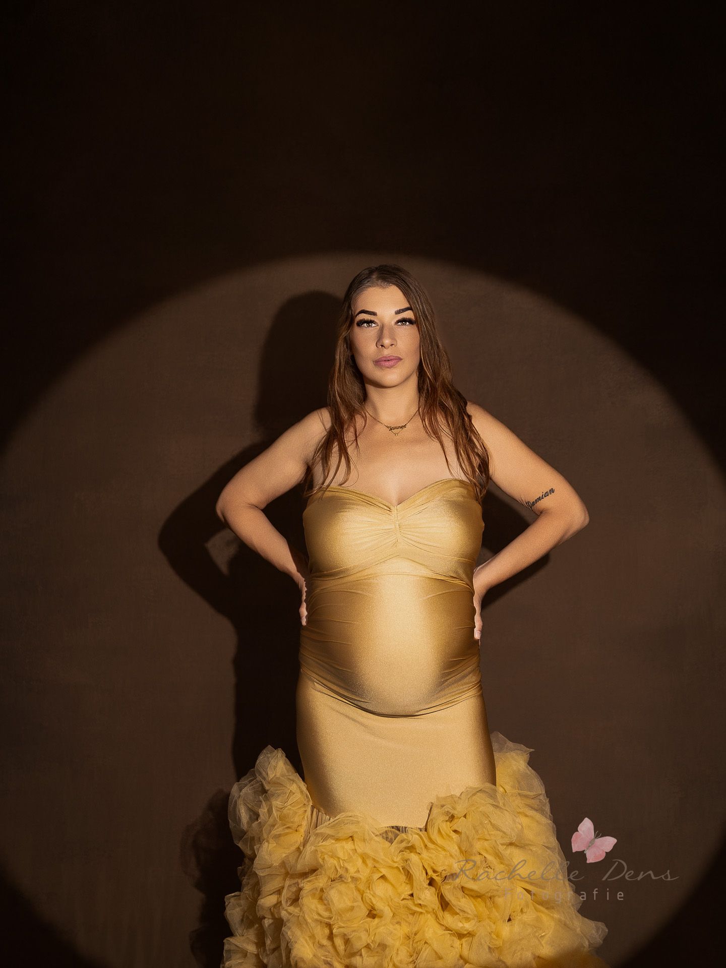 A person in a flowing gold evening gown poses dramatically against a dark background in a maternity photoshoot.