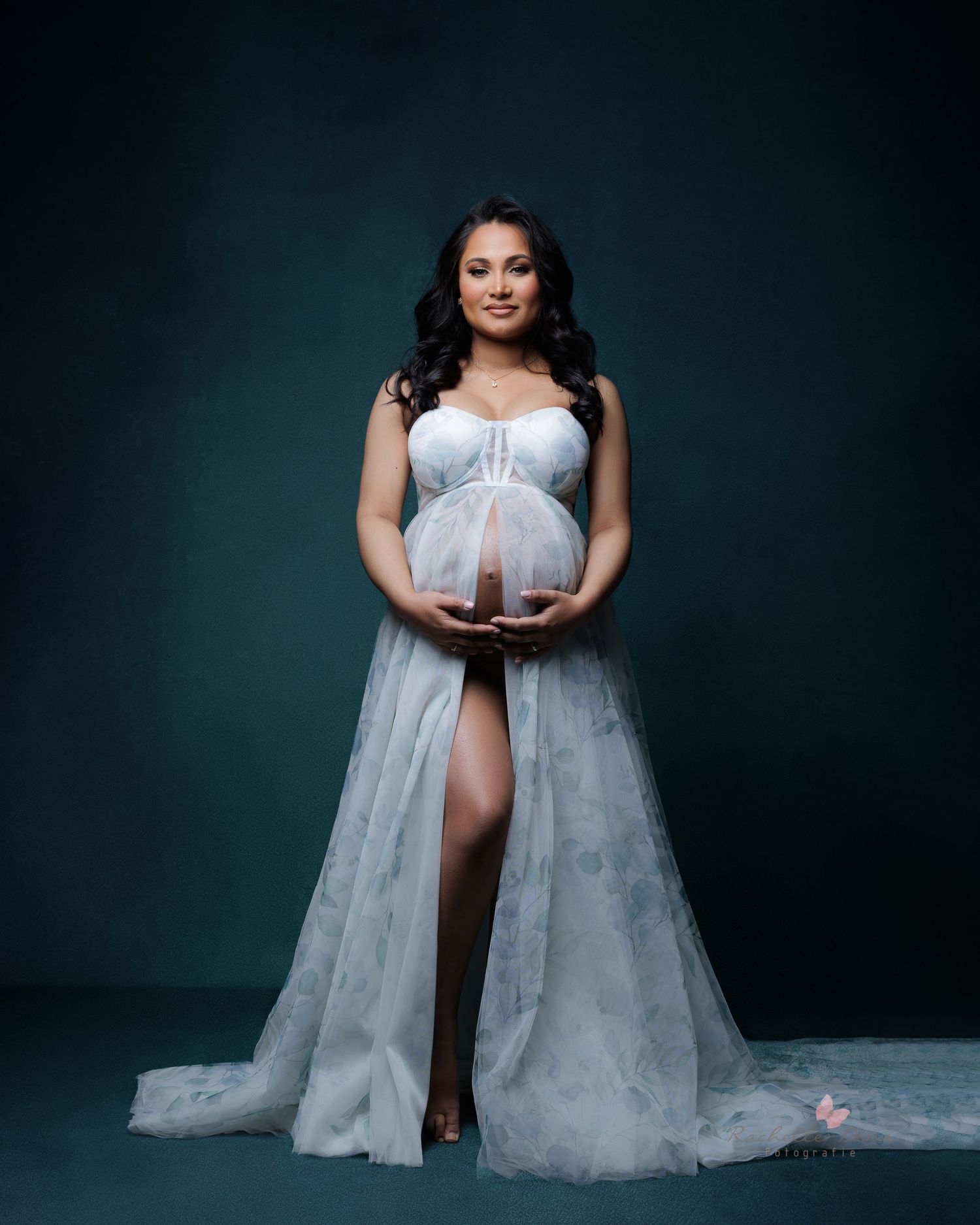 A maternity photo session featuring a flowing white tulle dress against a dark studio backdrop.