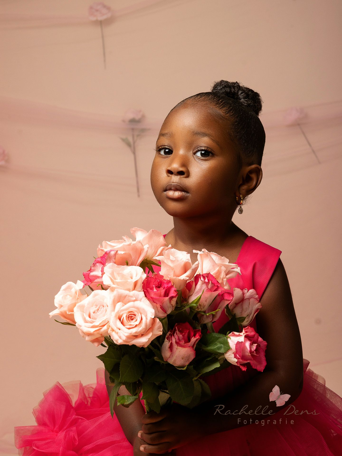 A child in a pink tulle dress poses with pink roses against a soft pink backdrop in a dreamy portrait series.