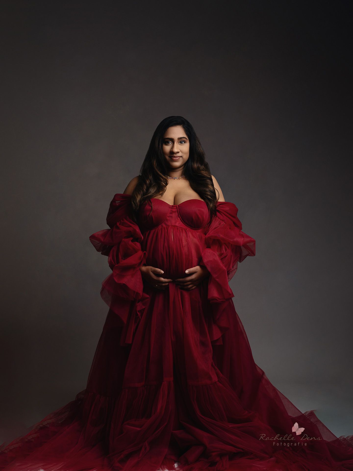 A dramatic burgundy tulle gown photographed in a moody studio setting against a dark background.