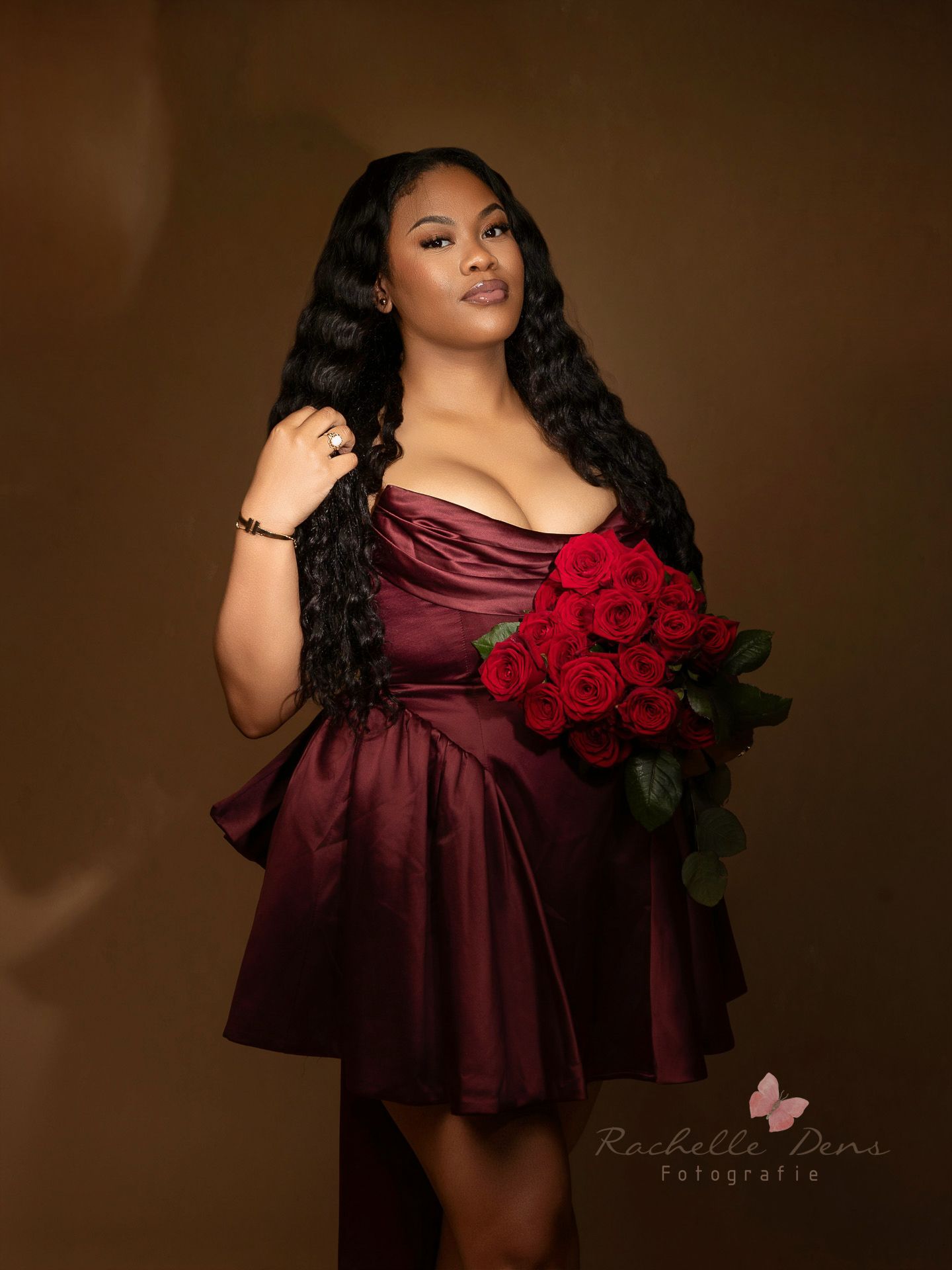 A person in a burgundy satin dress poses with red roses against a dark brown backdrop in a glamour photoshoot.