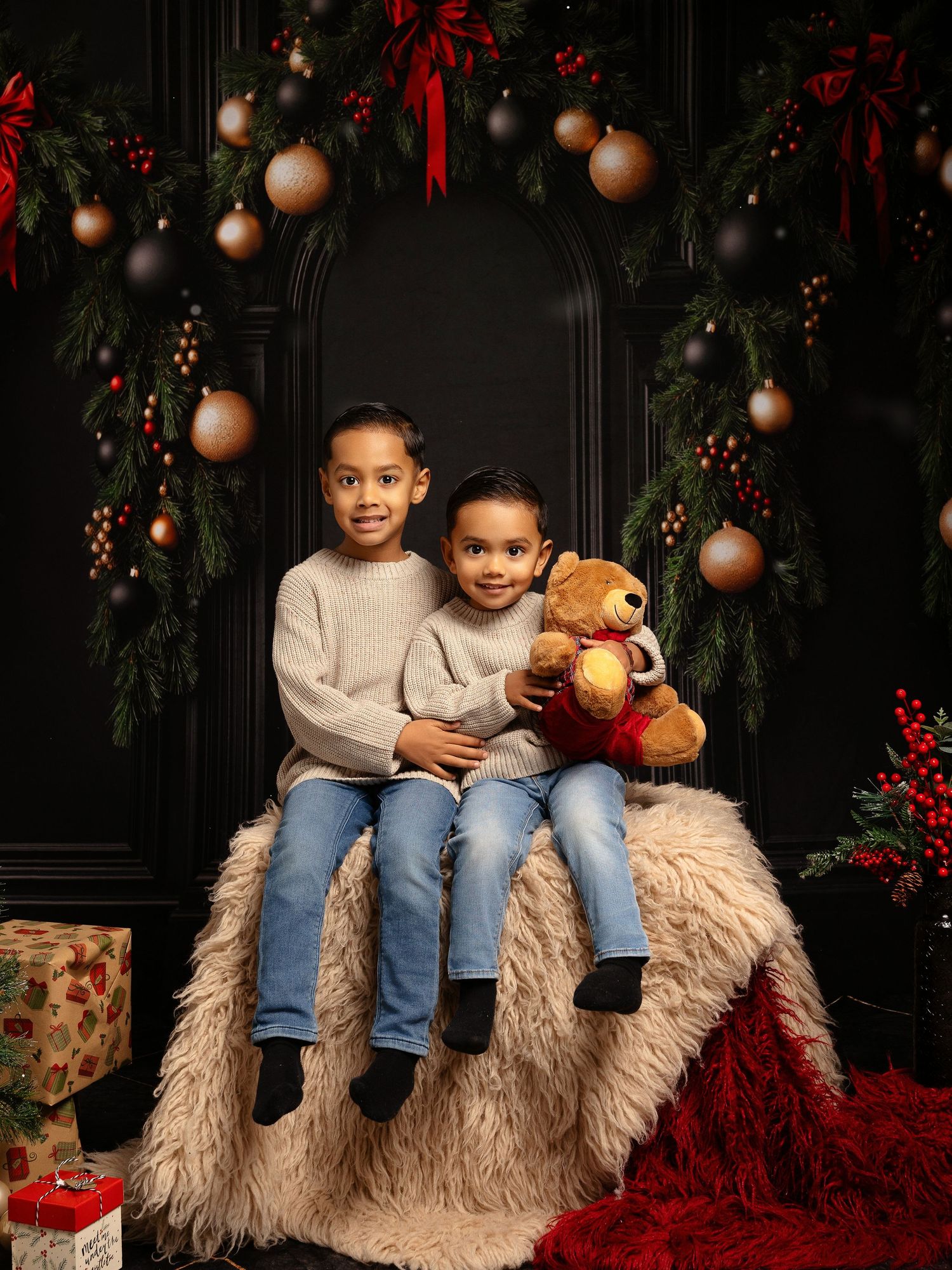 Two children in matching gray sweaters and jeans sit on a furry seat with a teddy bear amid Christmas decorations.