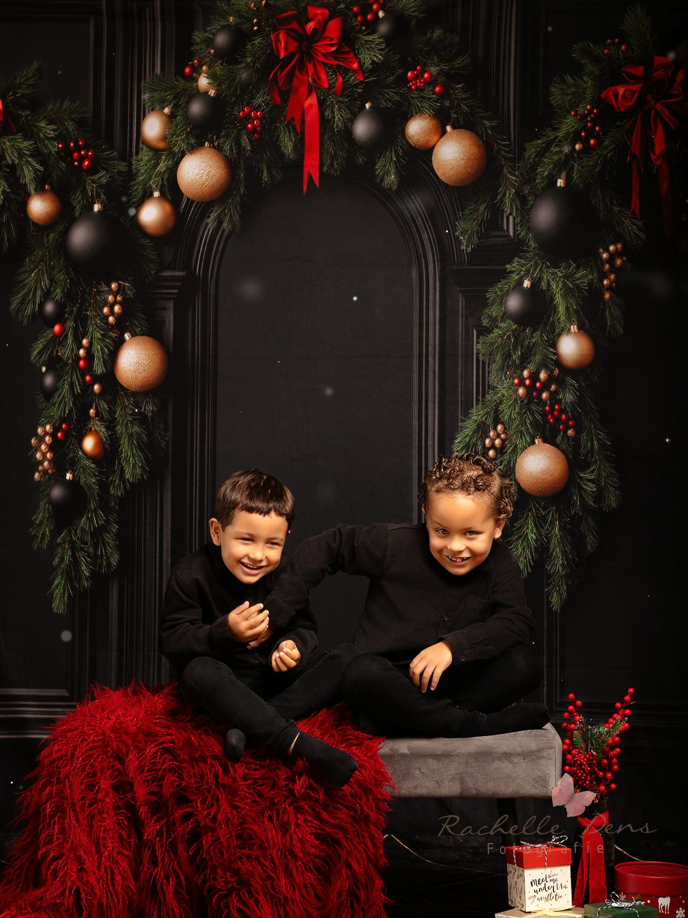 Two children in black outfits share a laugh while sitting together against a festive Christmas backdrop with ornaments.
