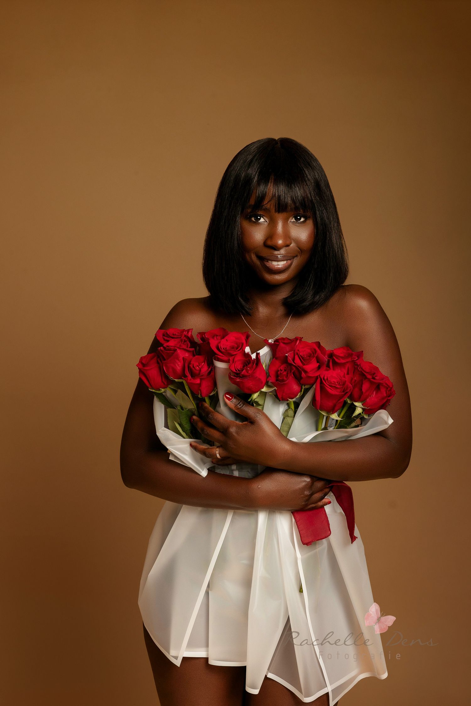 A person in a white dress holds a bouquet of vibrant red roses against a warm brown backdrop.