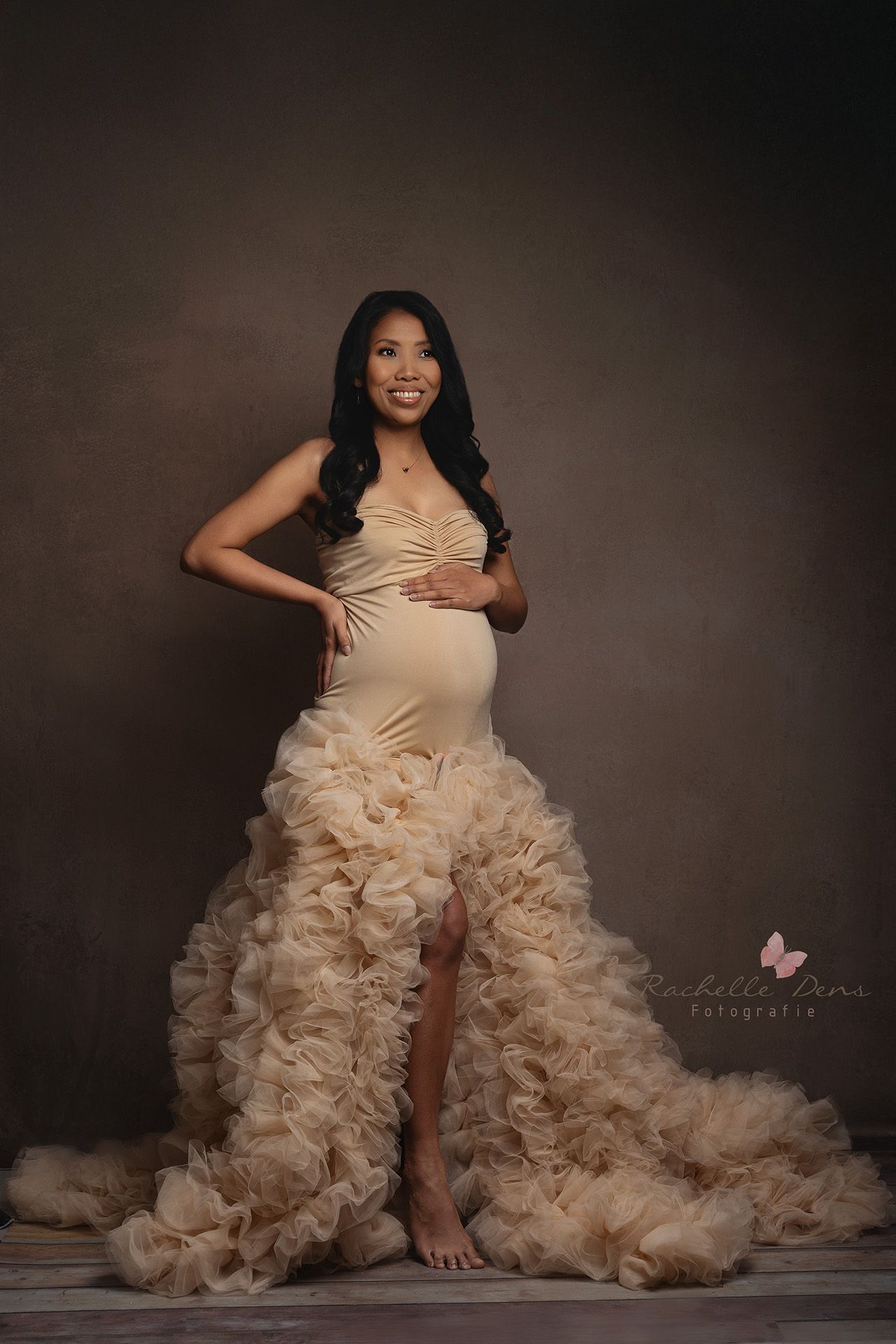Elegant maternity portrait in cream-colored ruffled gown with dramatic train against dark studio backdrop.