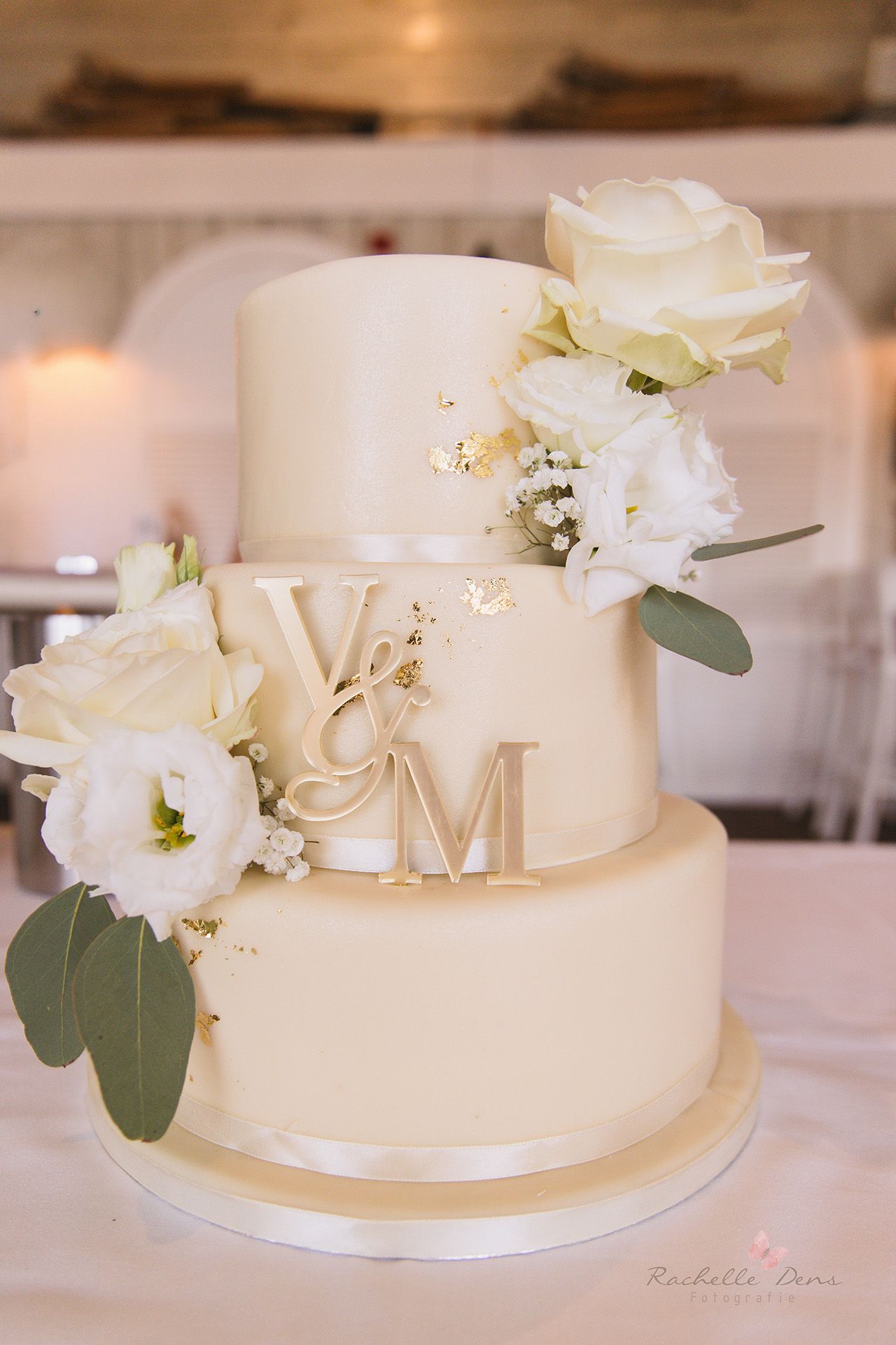 Elegant three-tier white wedding cake decorated with gold monogram letters and fresh white flowers and eucalyptus leaves.